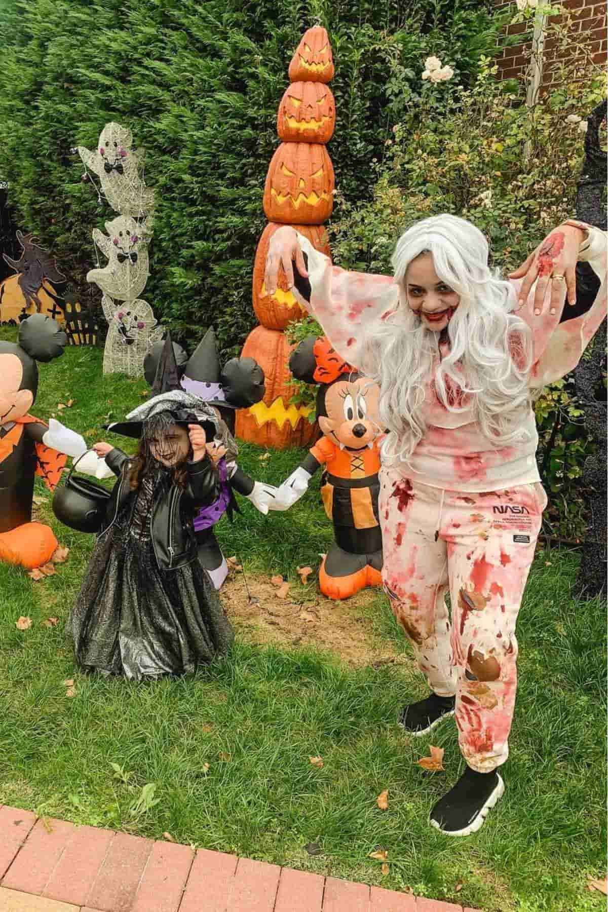 Two people in Halloween costumes pose outside; one is dressed as a witch and the other as a zombie, with inflatable pumpkin and cartoon decorations in the background.