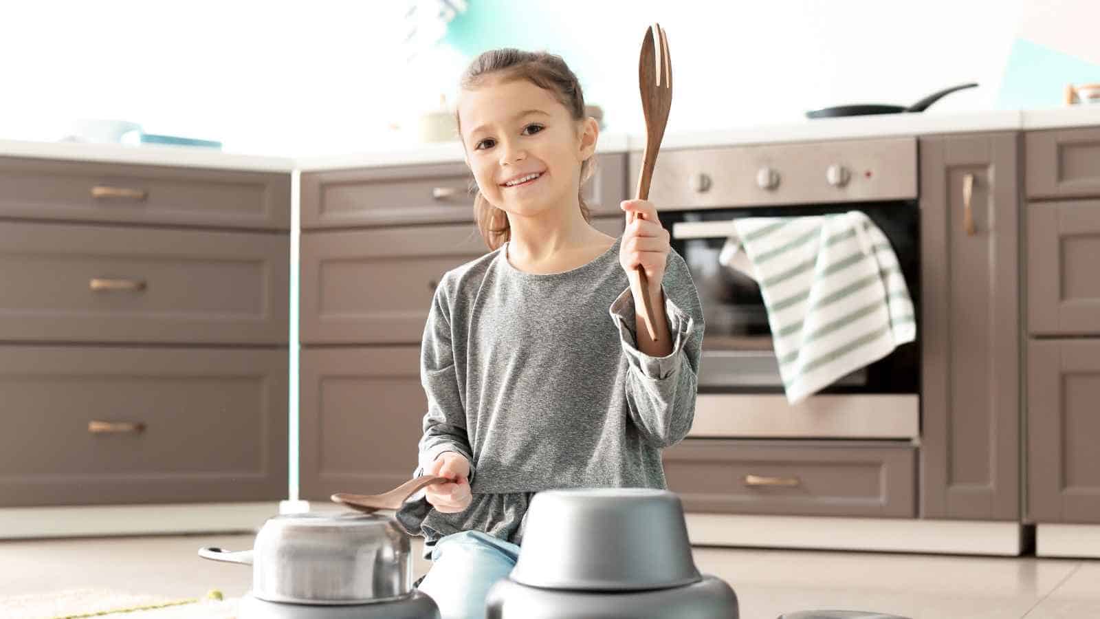 A young girl sits on a kitchen floor, smiling and holding wooden utensils, with several pots and pans arranged in front of her.