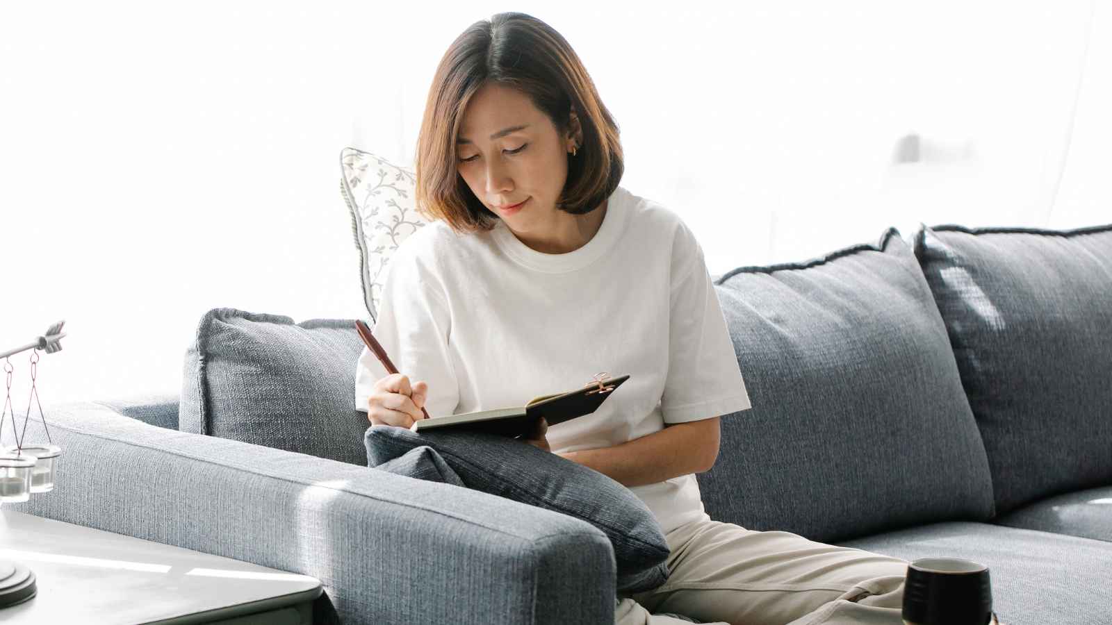 A woman sits on a grey sofa writing in a notebook, with a pen in hand and a coffee cup on the table beside her.