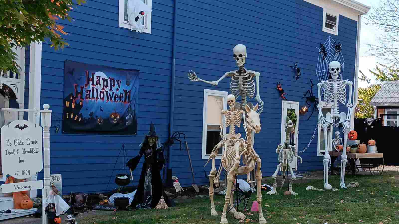 Halloween decorations on a blue house, including skeletons, a witch, spider webs, amazon halloween yard decorations, and a "Happy Halloween" banner. A white sign in front indicates vacancies at an inn.