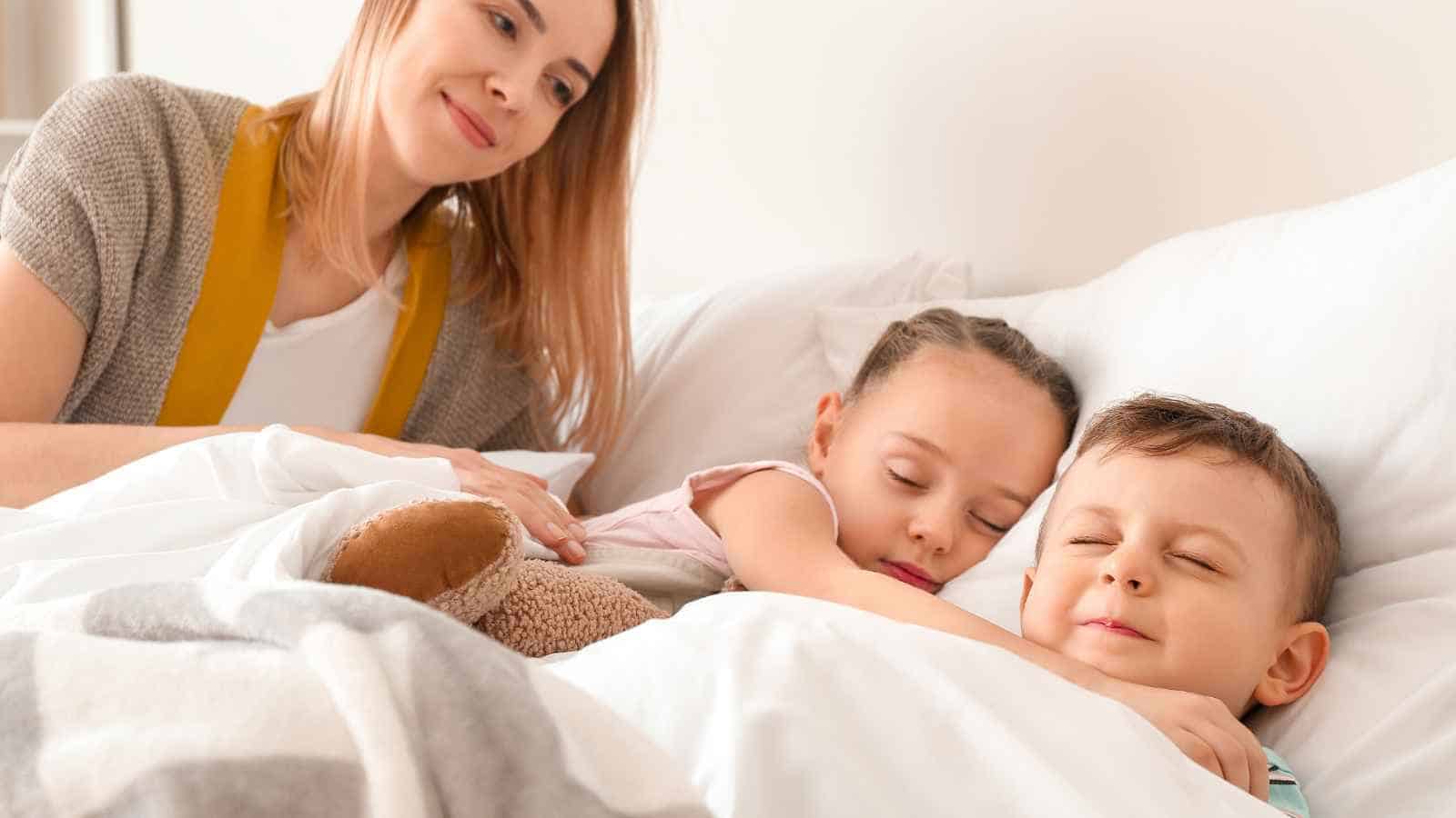 A woman sits beside two young children who are sleeping in bed, covered by a white blanket, with a stuffed animal nearby.