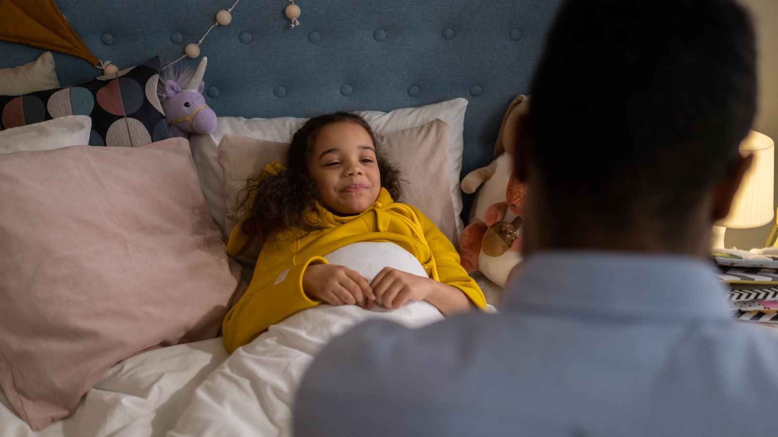 A child in a yellow hoodie lies in bed smiling, while an adult sits nearby with their back to the camera. Stuffed toys and pillows are on the bed.