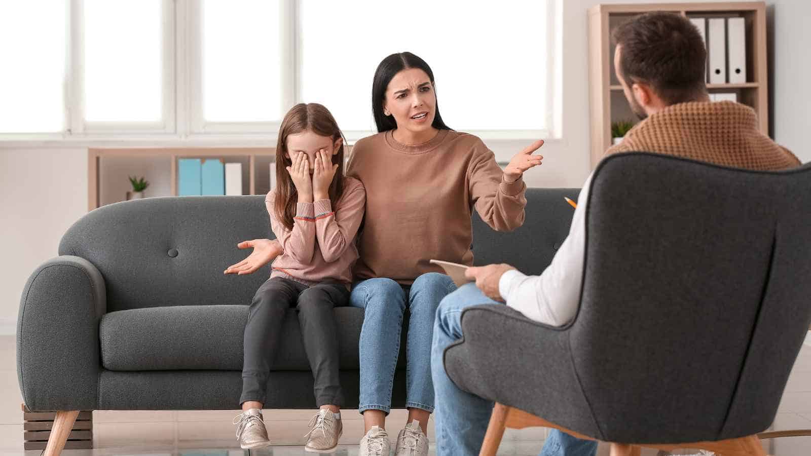 A woman and a girl sit on a couch; the girl covers her face while the woman gestures and talks to a man holding a clipboard, likely during a counseling session.
