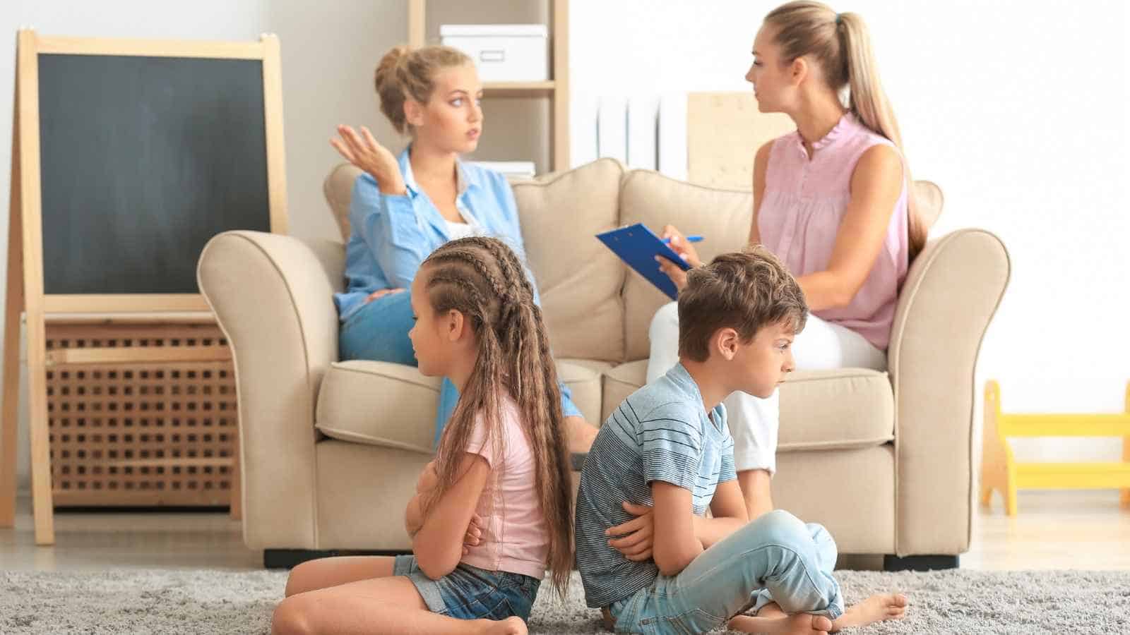 Two women sit on a couch having a discussion, while a girl and a boy sit on the floor in front of them with arms crossed, facing away from each other.