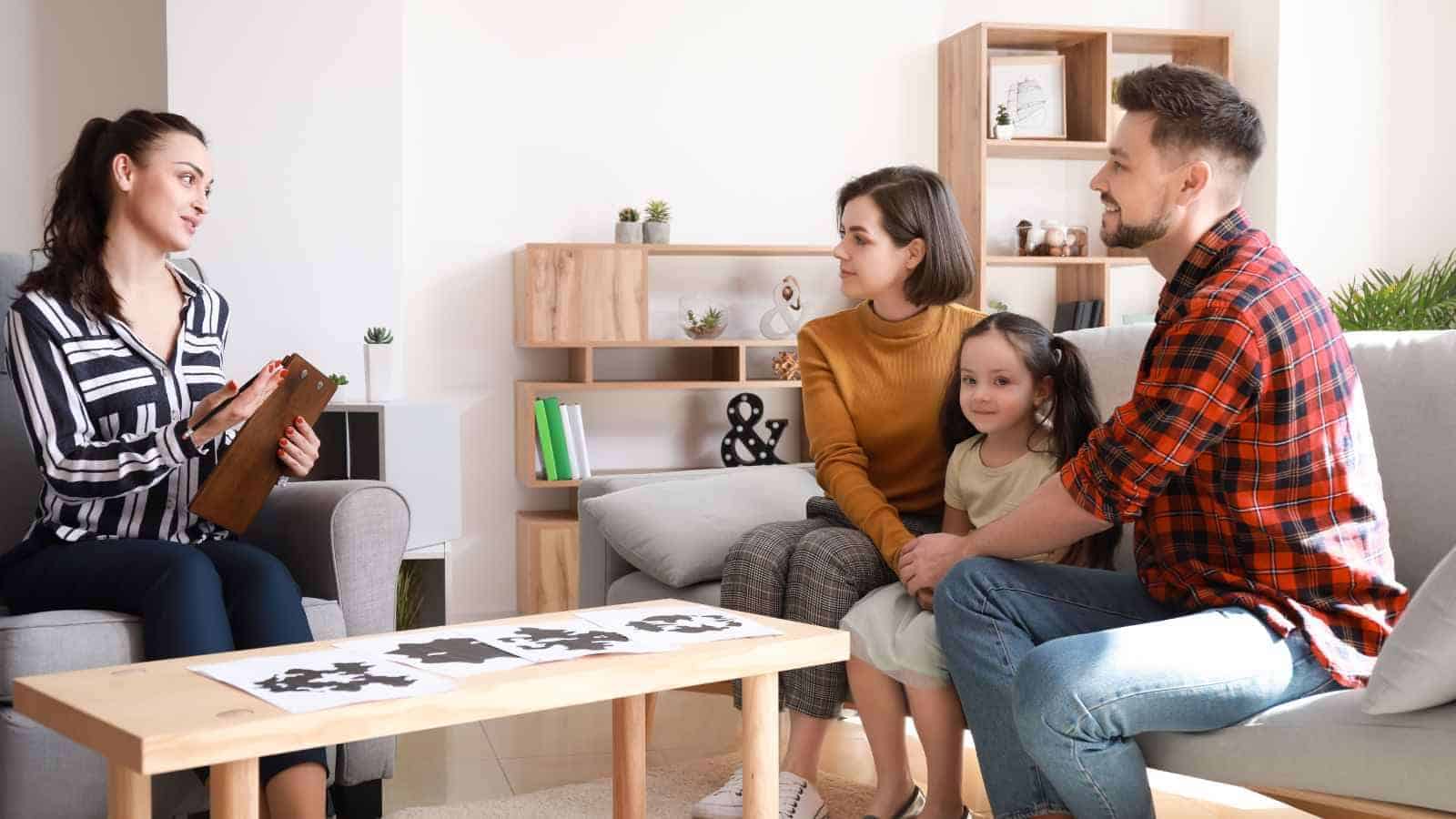 A woman with a clipboard talks to a couple and their young daughter sitting together on a couch in a modern living room.