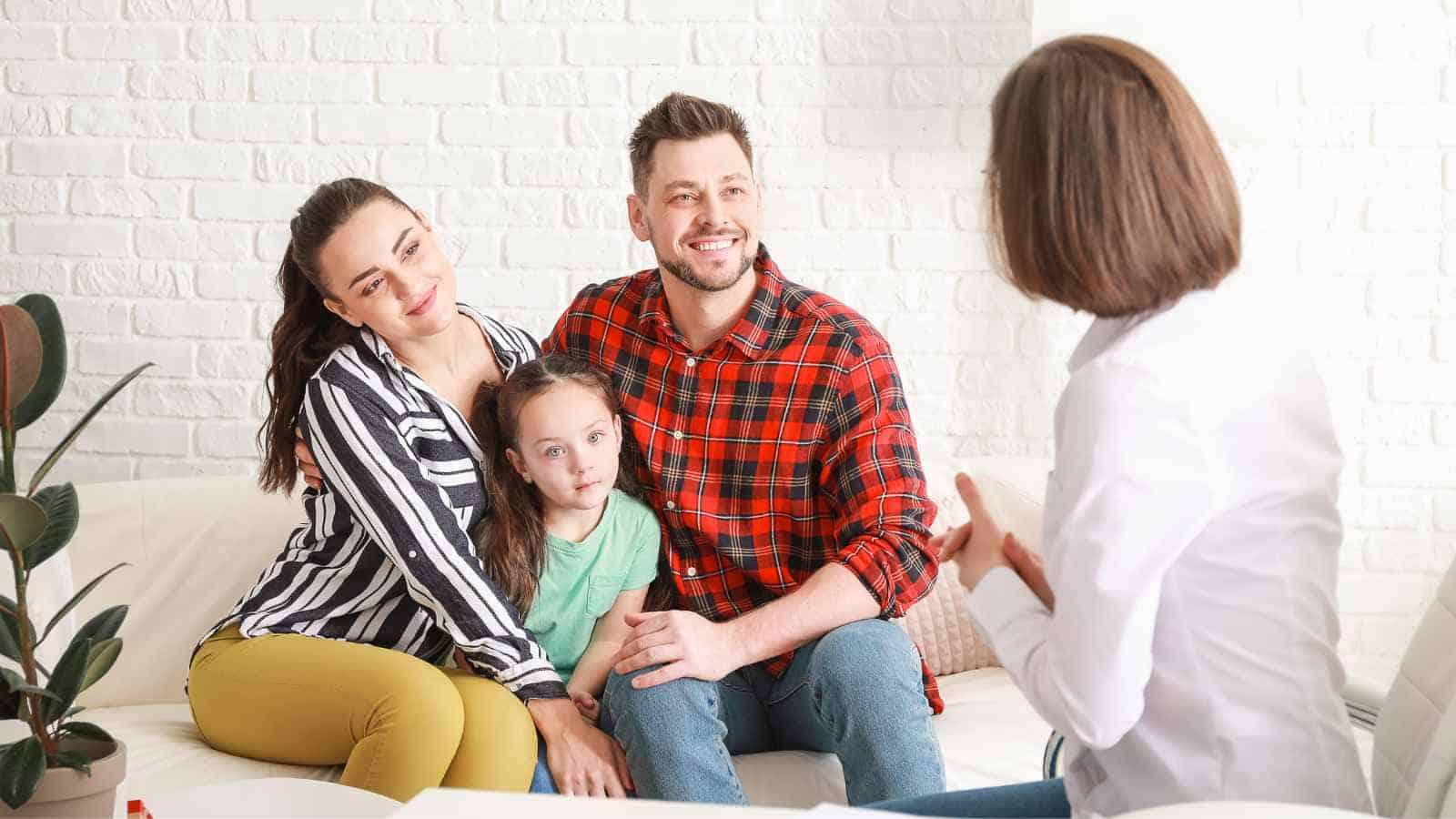 A couple and their young daughter sit on a couch, listening to a woman in a white coat who appears to be speaking to them in an office setting.