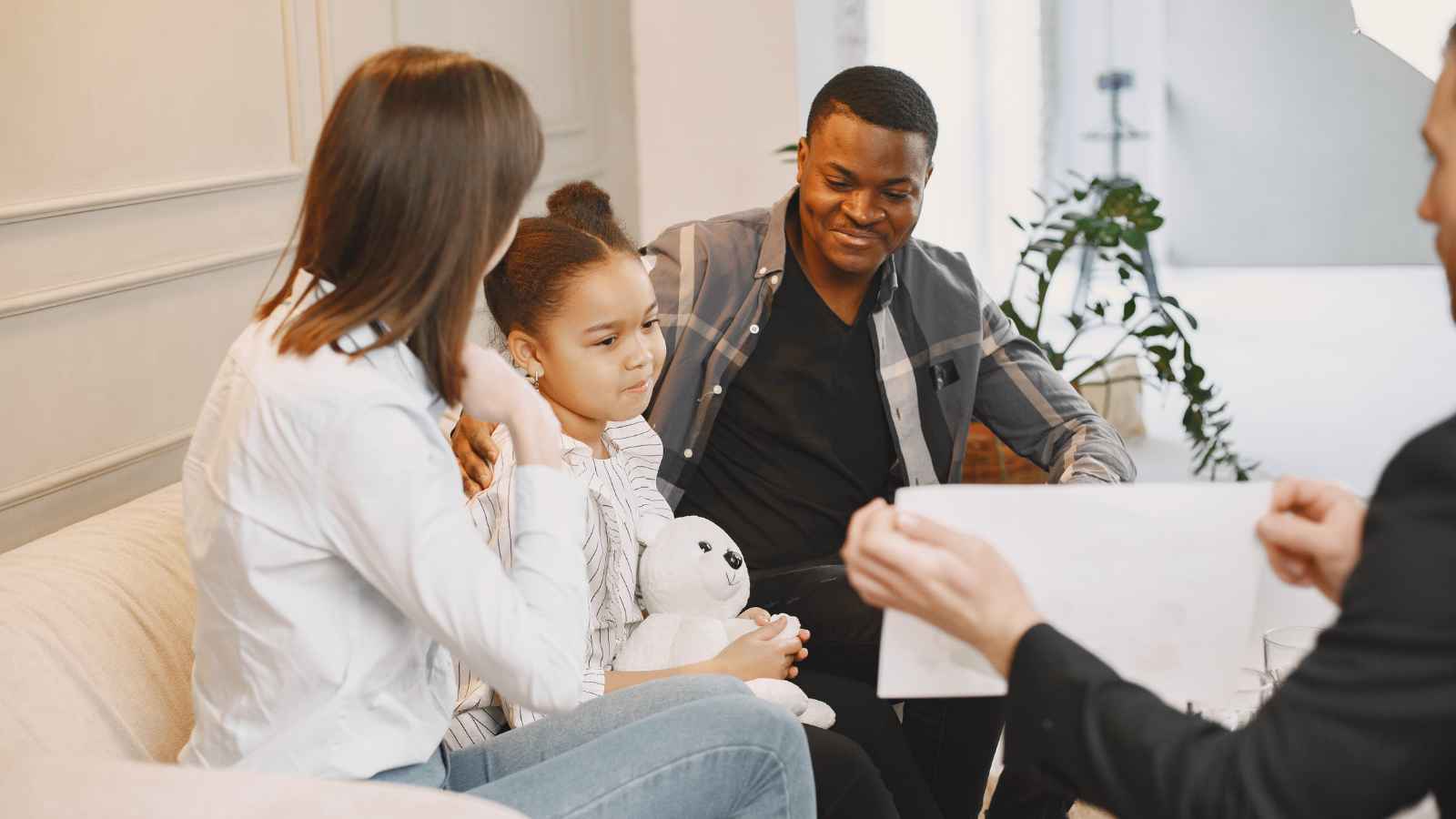 A family sits together on a couch while a man in a suit shows them a document; the child holds a stuffed animal.