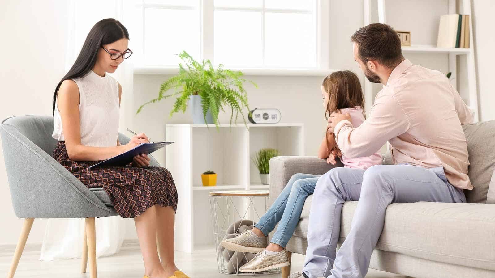 A woman with a clipboard speaks to a man and a young girl sitting on a couch in a bright, modern office.