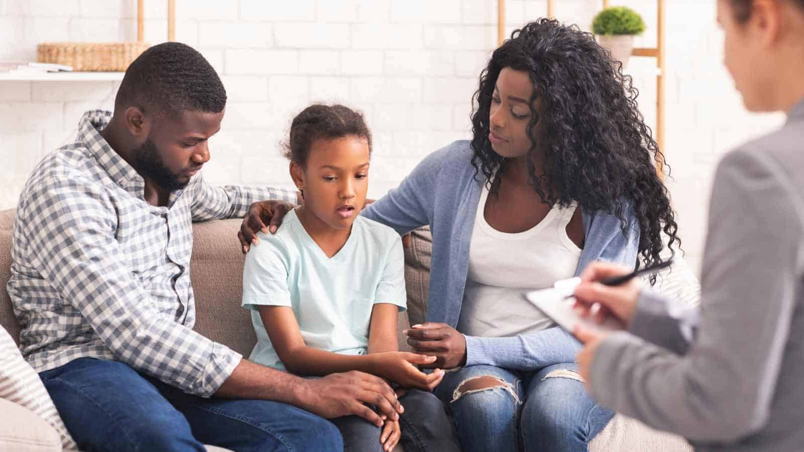A child sits between two adults on a couch, looking down, while an adult takes notes nearby, suggesting a counseling or therapy session.