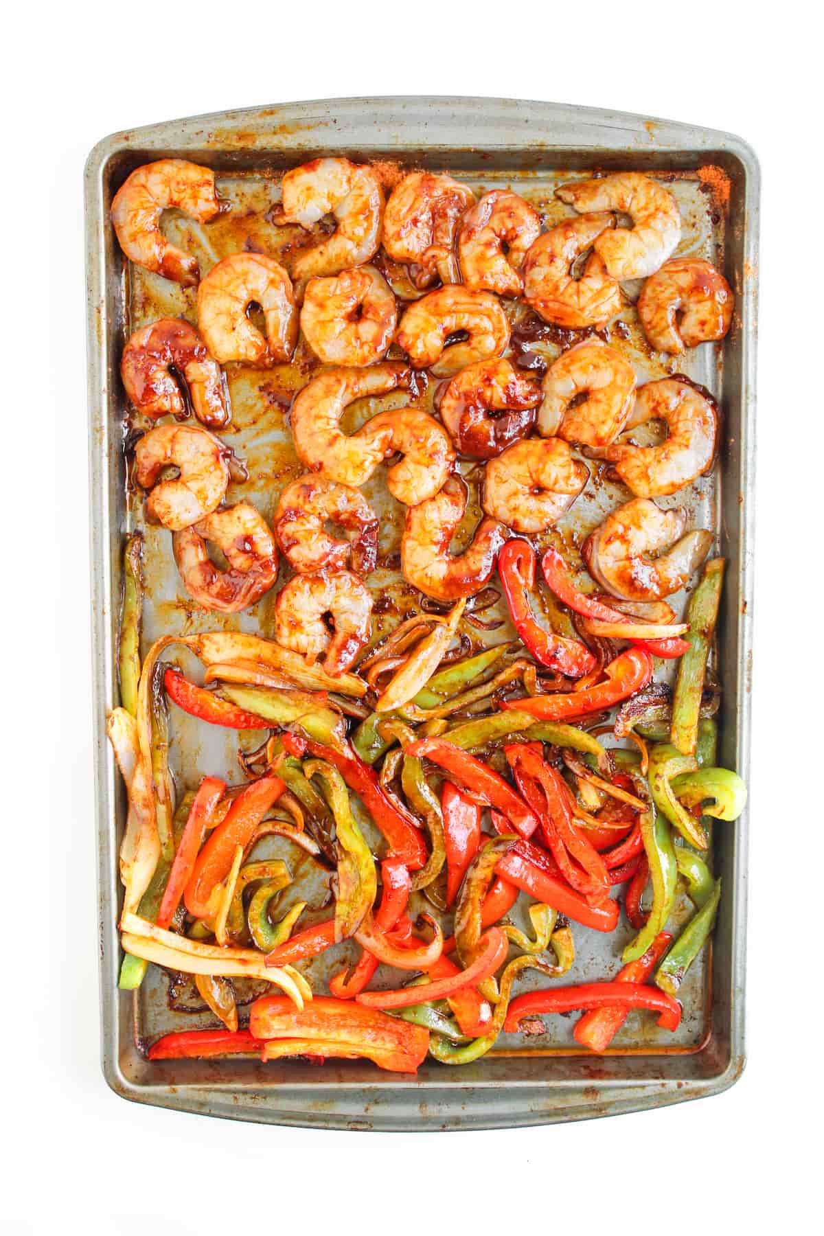 Sheet pan with cooked shrimp on the top half and roasted red, yellow, and green bell pepper strips on the bottom half, on a white background.