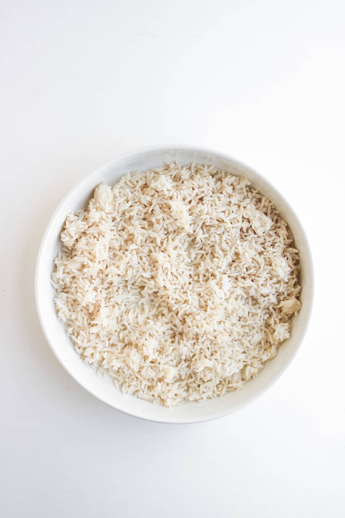A white bowl filled with cooked white rice, placed on a white surface.