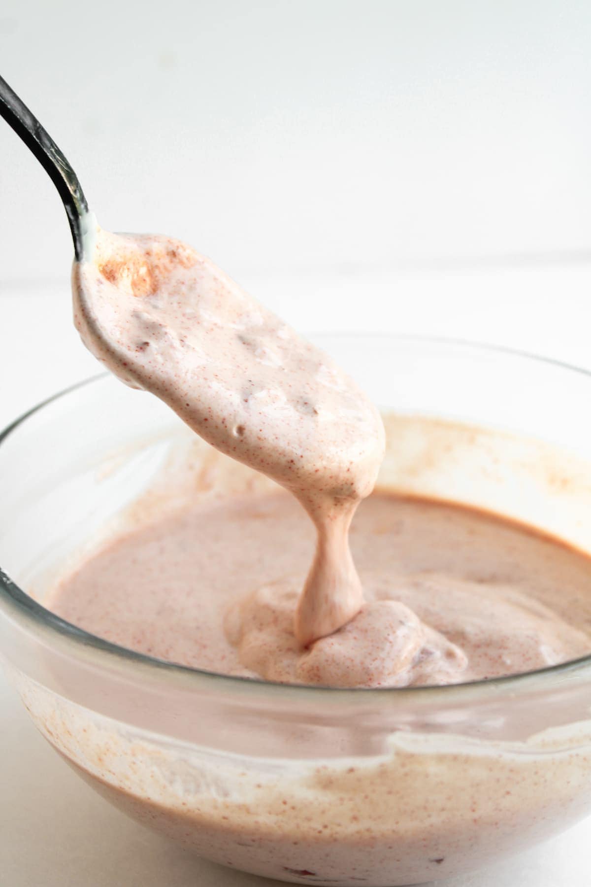 A spoon lifts thick, creamy pink mixture from a glass bowl against a plain white background.