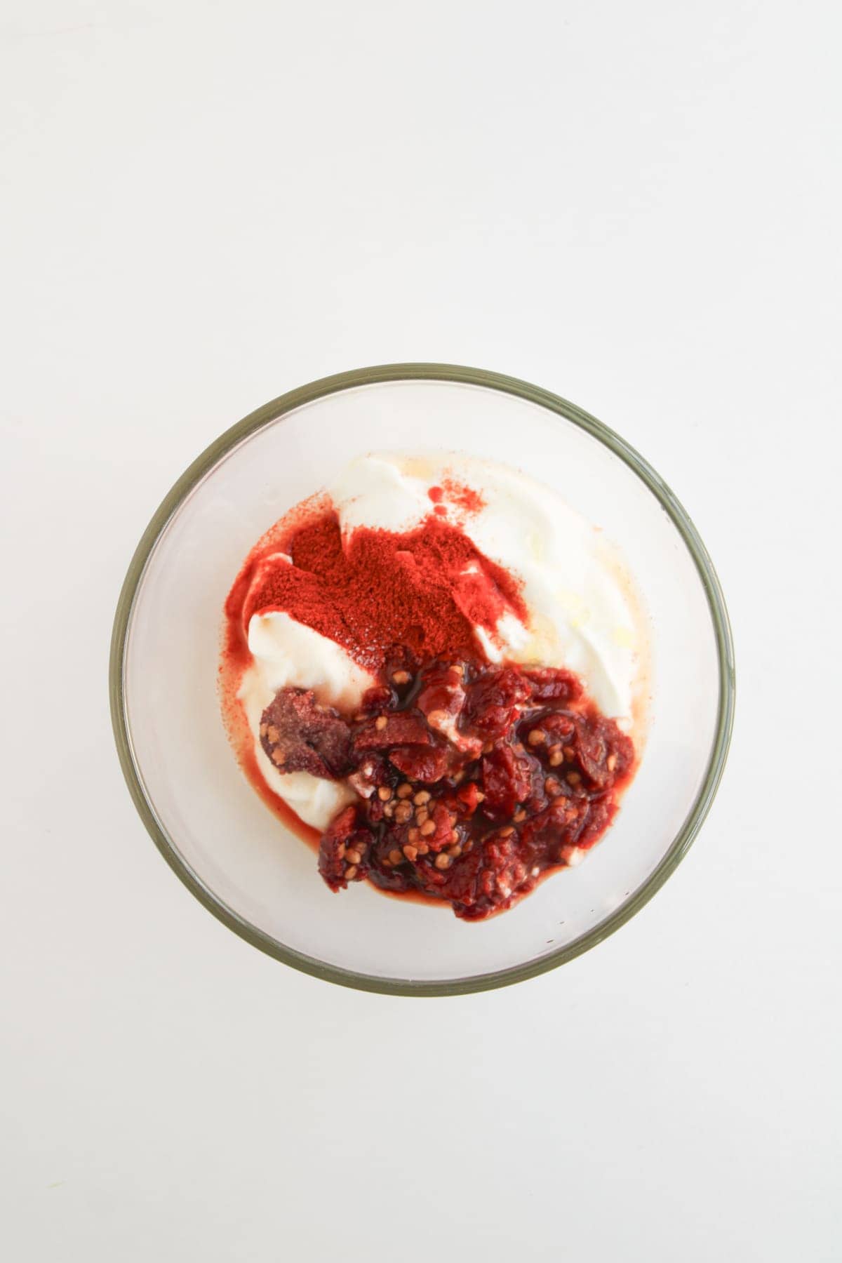 A clear glass bowl with yogurt, a scoop of berry compote, and a sprinkle of red powder on a white surface.