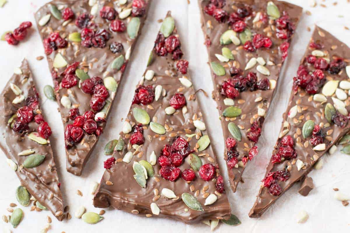Chocolate bark pieces topped with dried cranberries, pumpkin seeds, sunflower seeds, and flax seeds on a white surface.