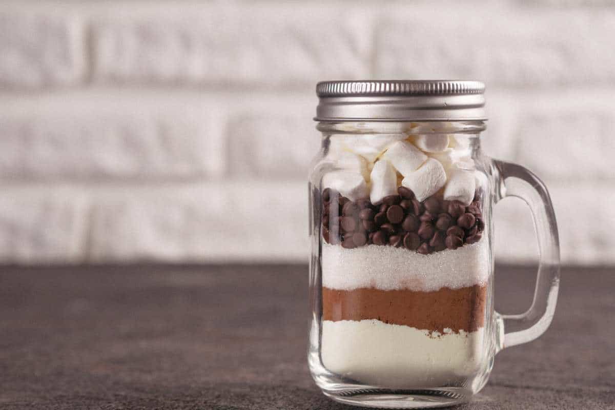 A mason jar filled with layered dry ingredients for hot chocolate, including cocoa powder, sugar, chocolate chips, and mini marshmallows, sits on a dark surface.