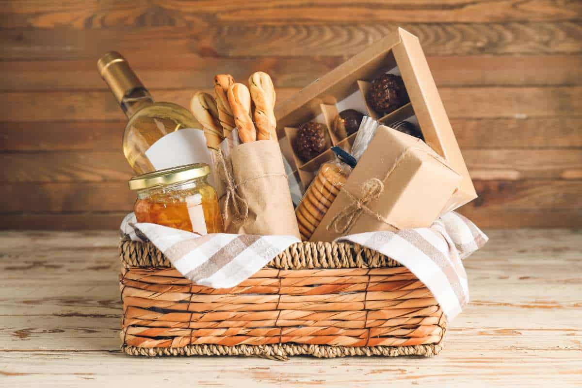 A wicker basket filled with a wine bottle, breadsticks, a jar of preserves, cookies, chocolates, and gifts, all arranged on a wooden surface with a checkered cloth.