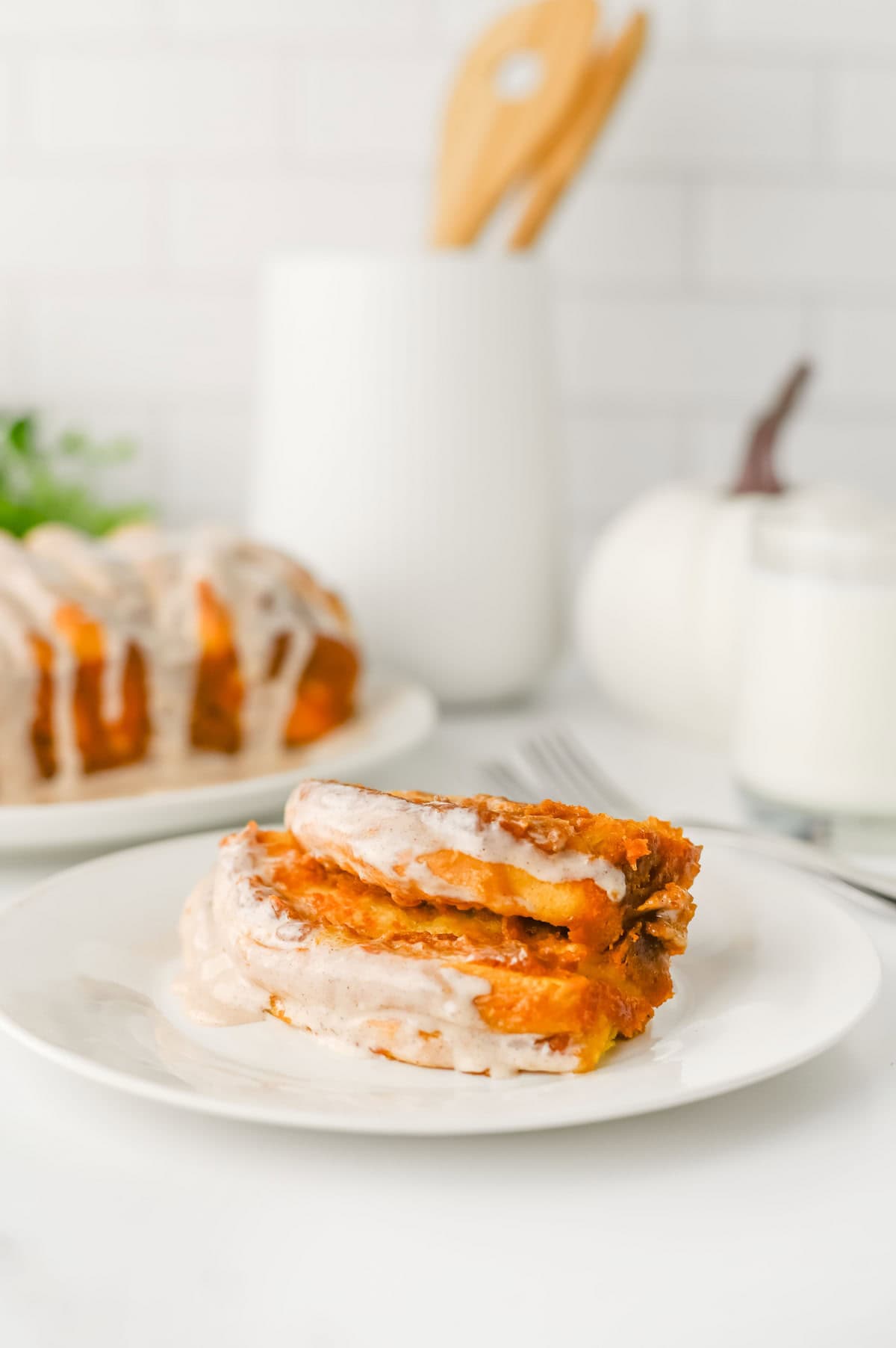 Two slices of pumpkin bread are on a white plate in the foreground, with a loaf and a glass of milk in the background on a white countertop.