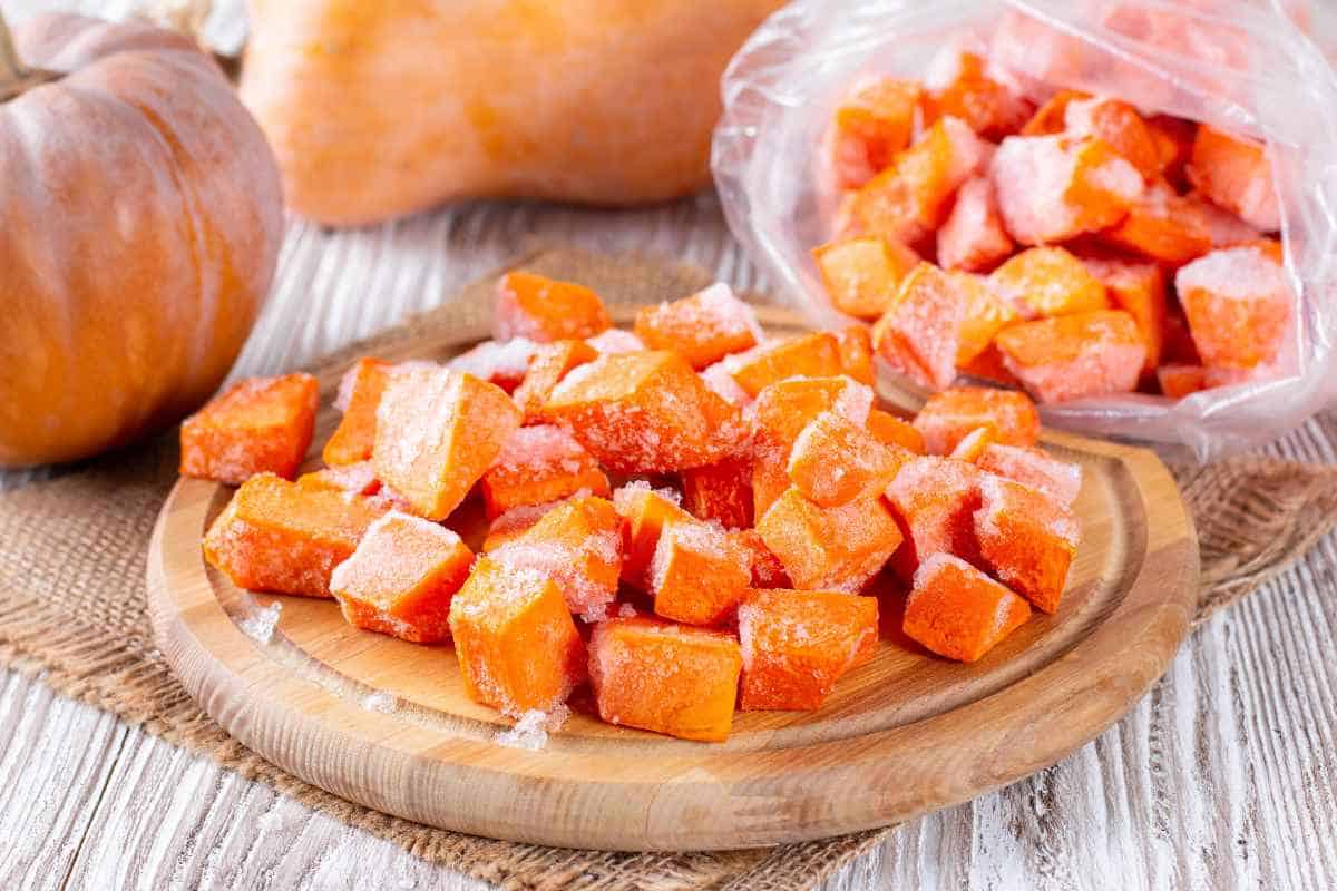 Frozen cubes of pumpkin, perfect for those learning how to freeze pumpkin, are arranged on a wooden plate, with extra cubes stored in a plastic bag in the background on a rustic table.