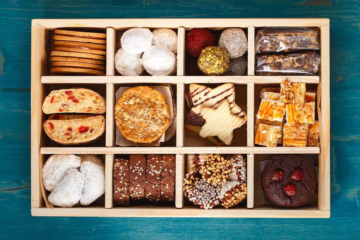 A wooden box with assorted cookies, biscuits, and sweets arranged in compartments on a blue surface.