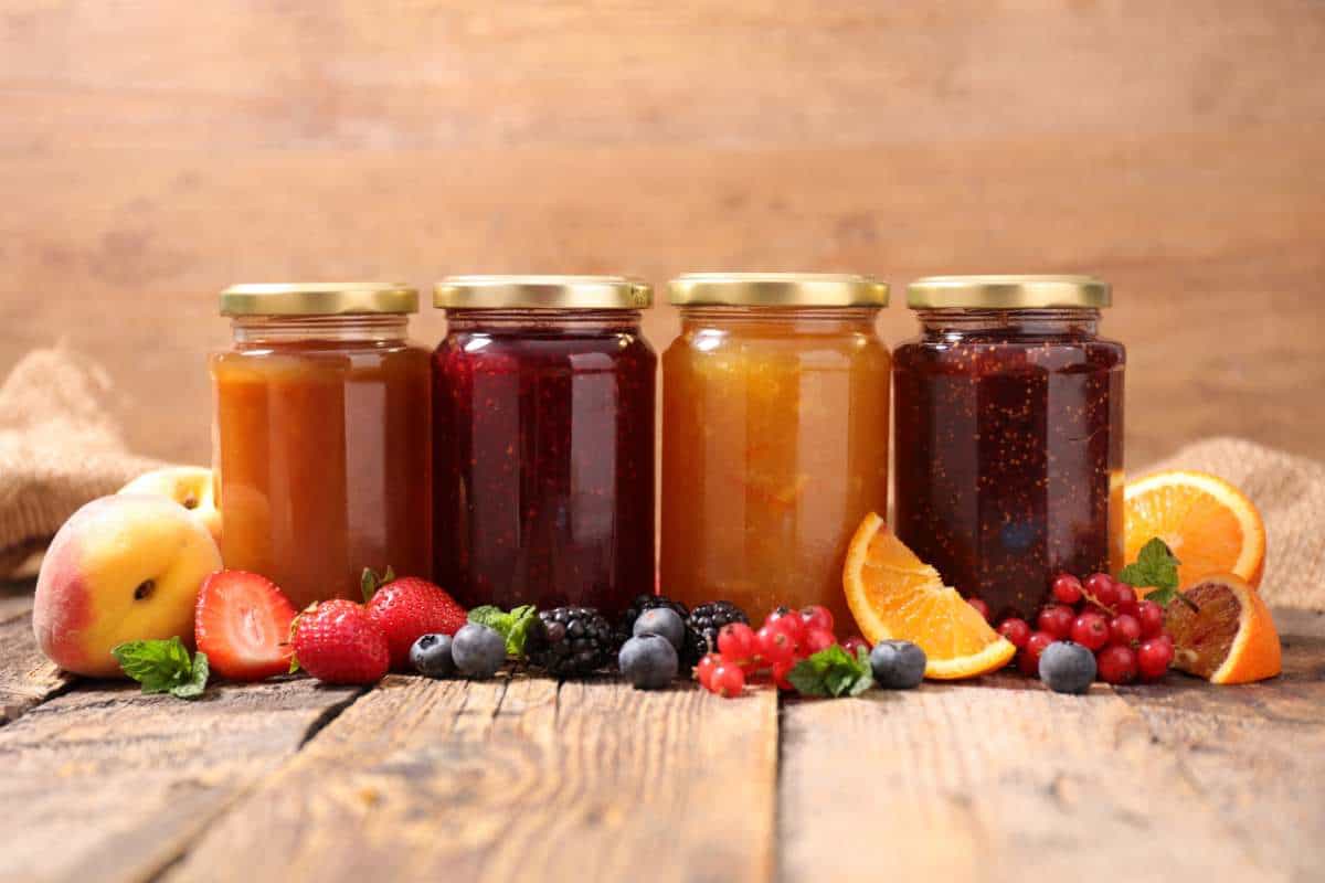 Four jars of assorted fruit jams with gold lids are lined up on a rustic wooden table, surrounded by fresh fruits including strawberries, peaches, blueberries, oranges, and red currants.