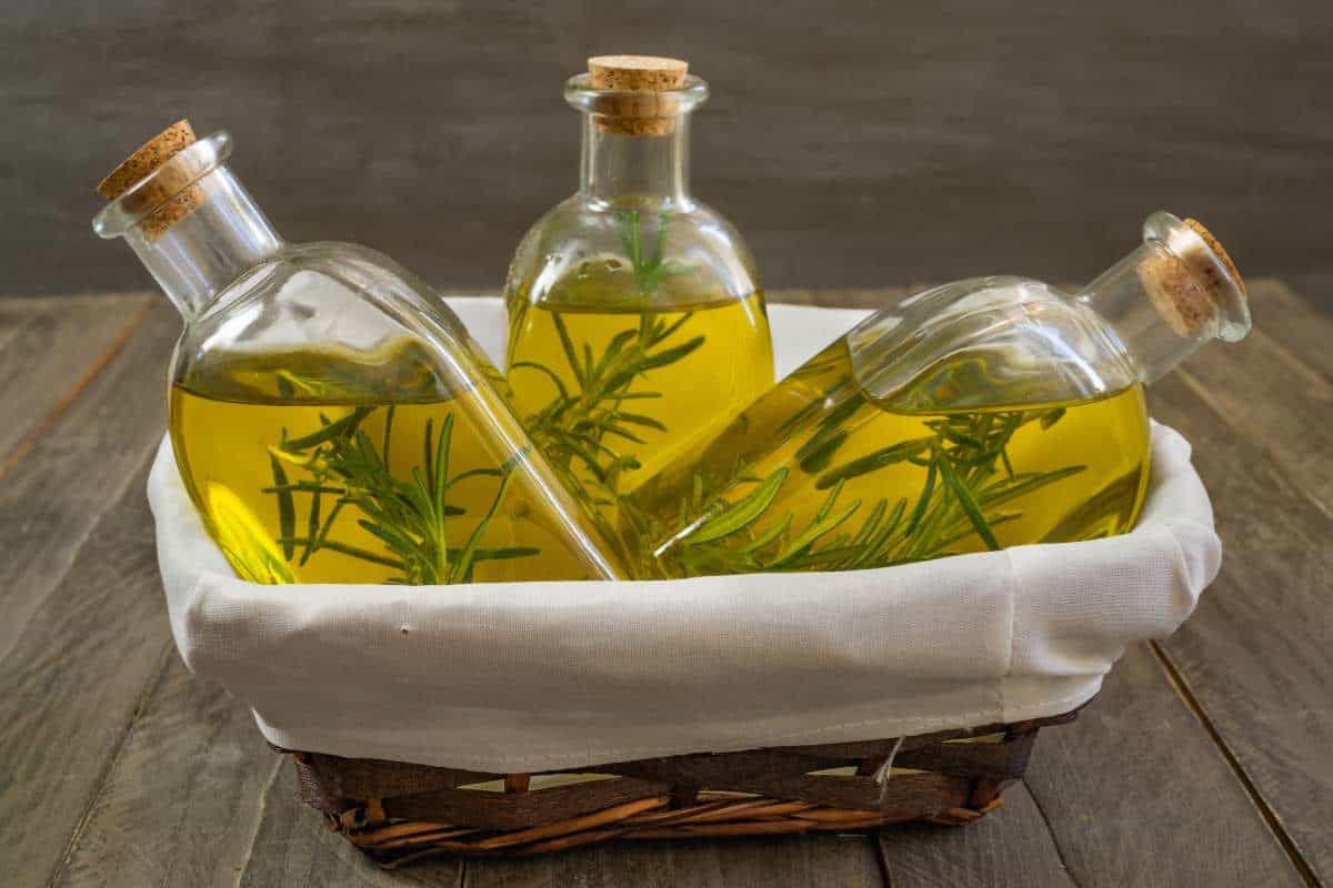 Three glass bottles filled with olive oil and sprigs of rosemary sit in a lined wicker basket on a wooden surface.