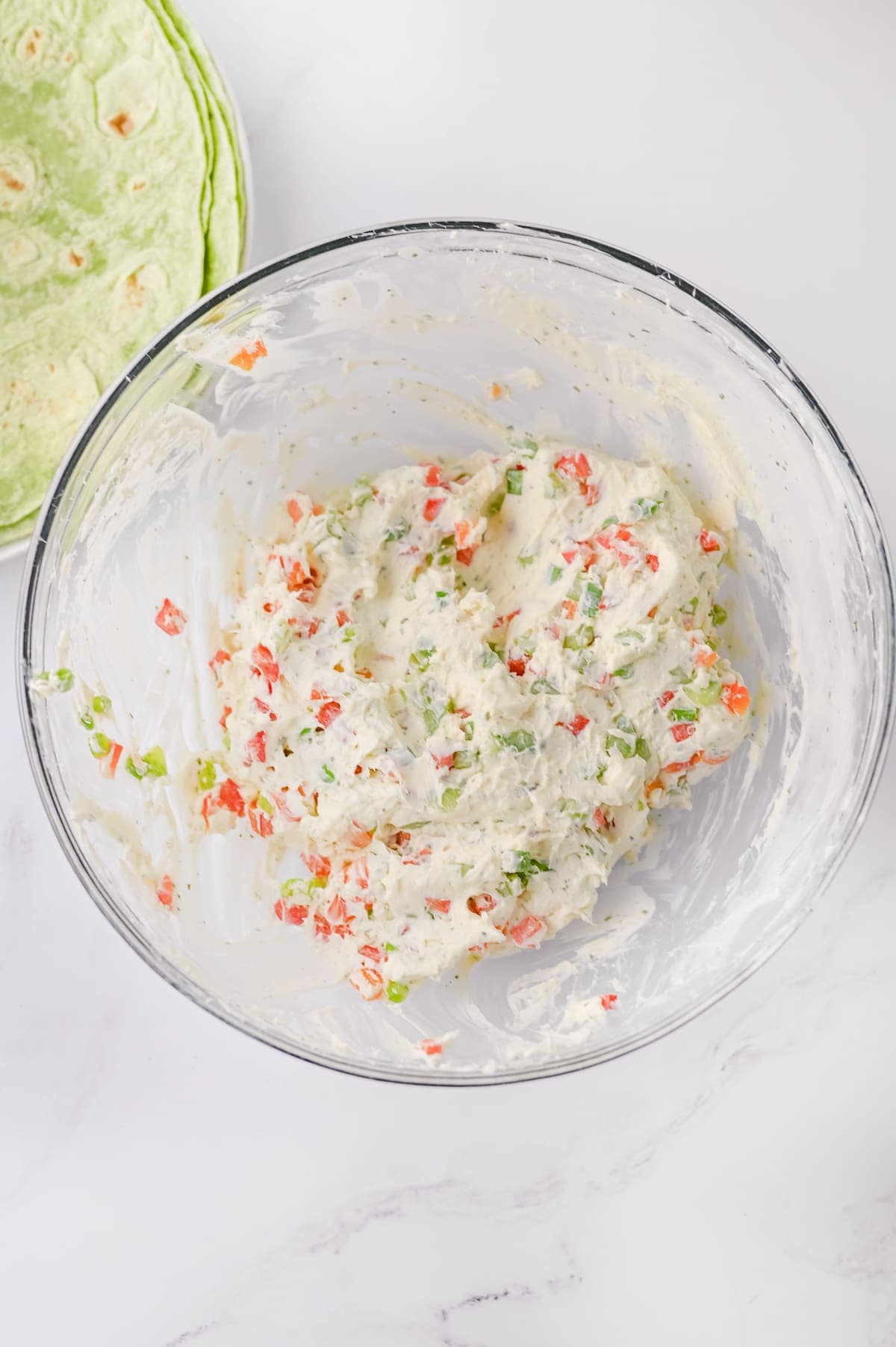 A glass bowl filled with a creamy mixture containing chopped red and green peppers, with green tortillas partially visible in the background.