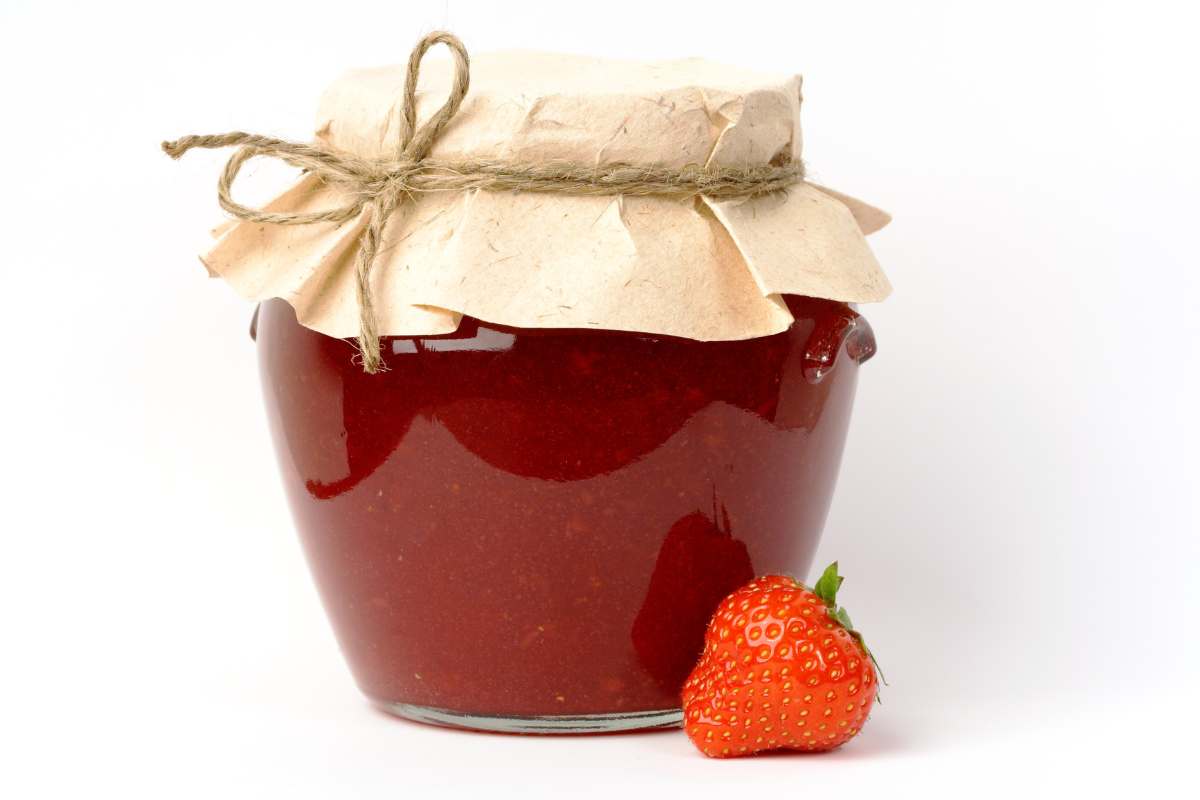 A glass jar filled with strawberry jam, covered with paper and tied with twine, sits next to a fresh strawberry on a white background.