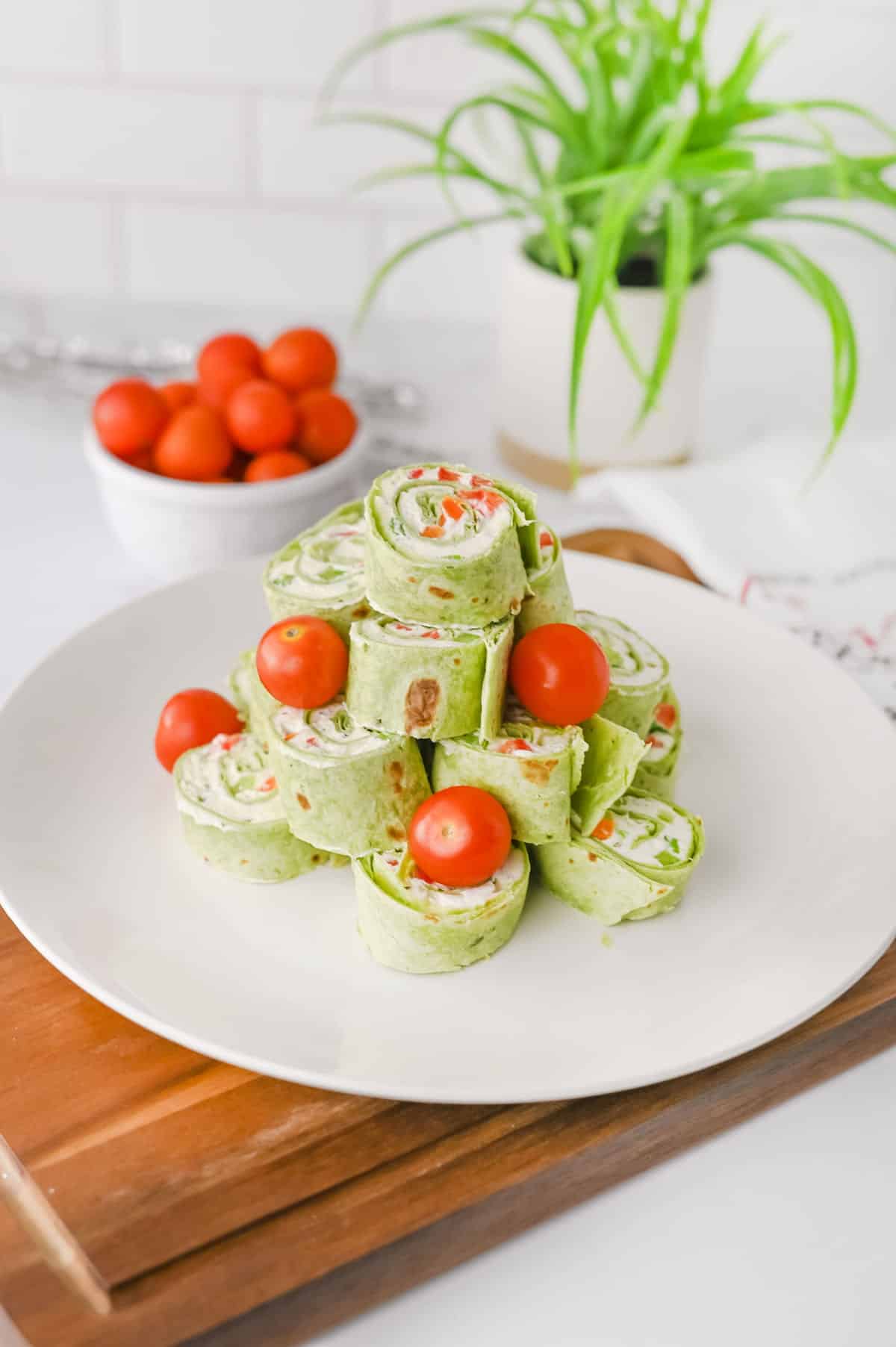 A white plate with green tortilla pinwheels filled with cream cheese and vegetables, garnished with whole cherry tomatoes, on a wooden board with a plant and bowl of tomatoes in the background.