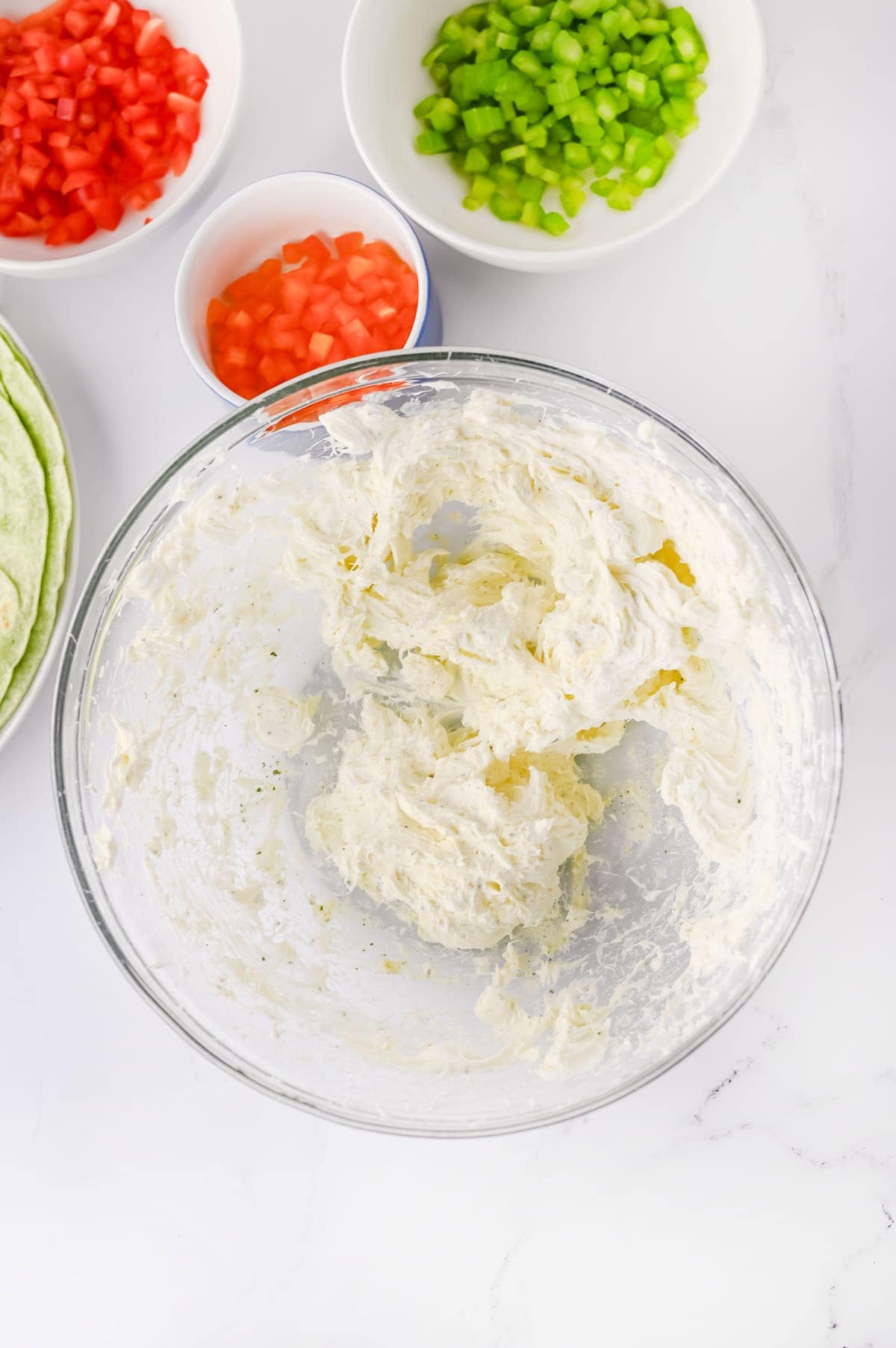 A glass bowl with a creamy mixture, surrounded by bowls of chopped green bell peppers and tomatoes on a white surface.