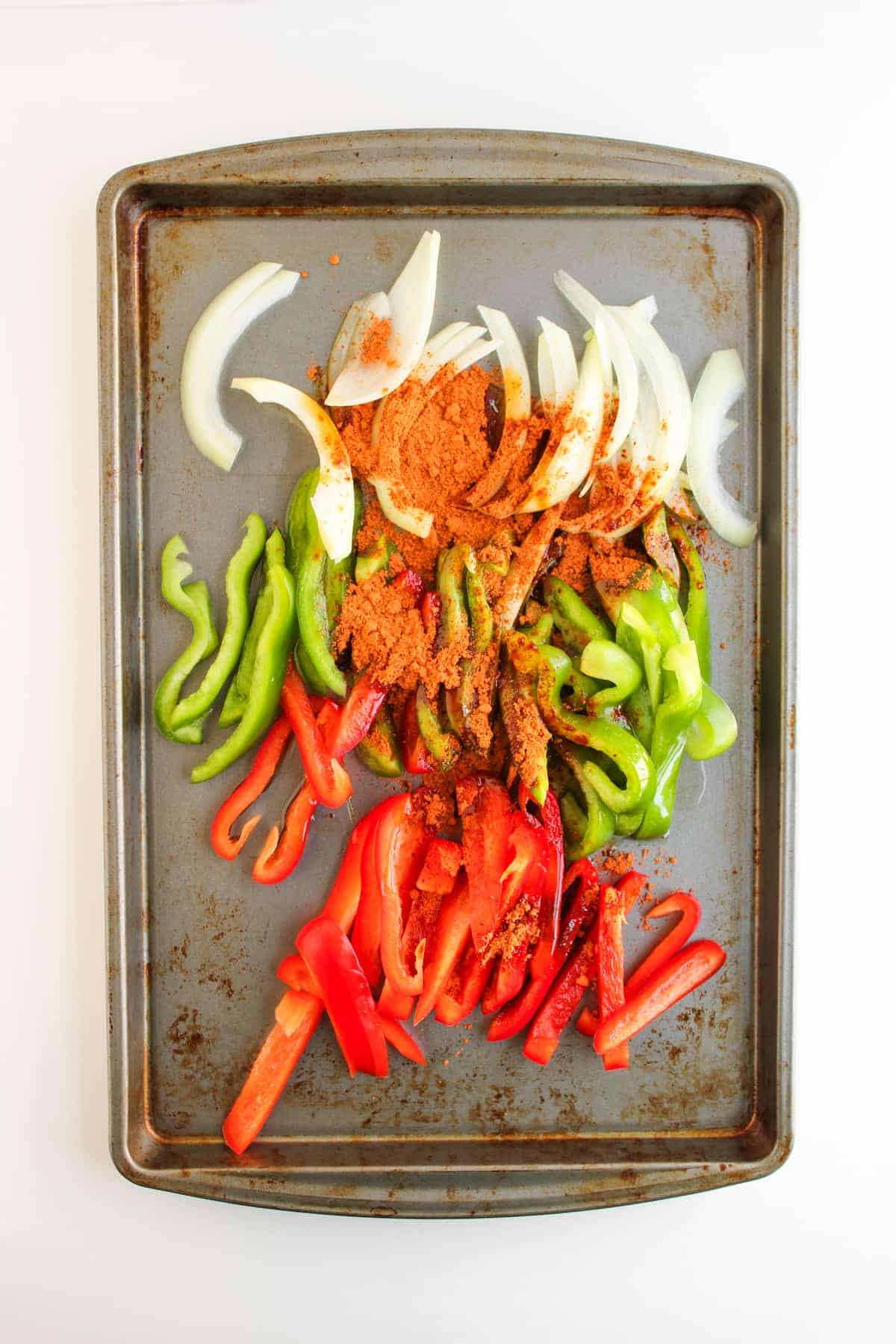 Sliced red and green bell peppers and onions with seasoning on a metal baking sheet, ready to be cooked.