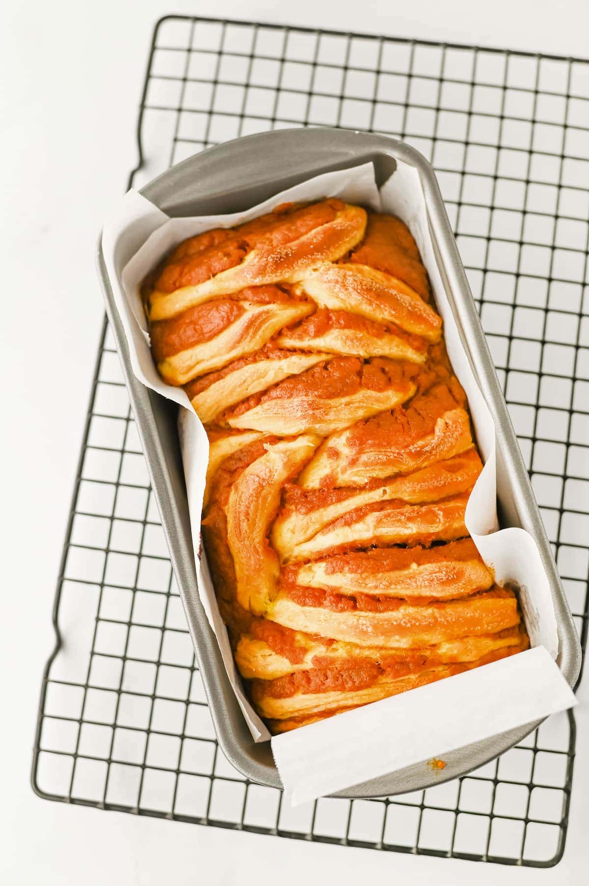 A freshly baked pumpkin bread with twisted layers sits in a parchment-lined loaf pan on a cooling rack.