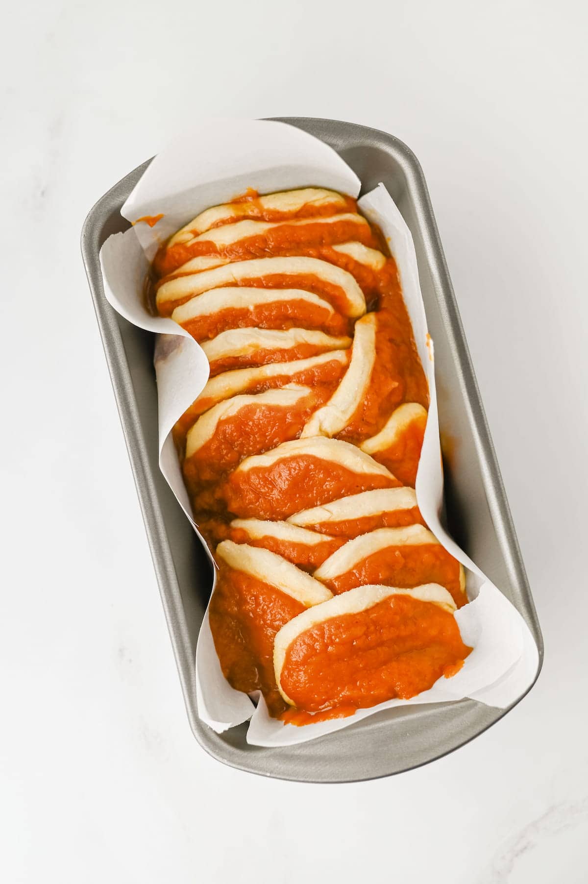 A loaf pan lined with parchment paper holds unbaked bread dough layered with tomato sauce.