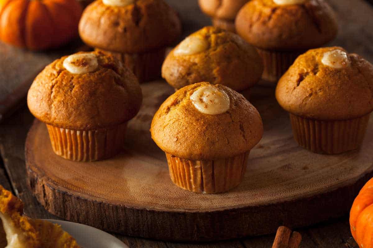 Pumpkin muffins with cream cheese centers are arranged on a wooden serving board, with small pumpkins and cinnamon sticks in the background.