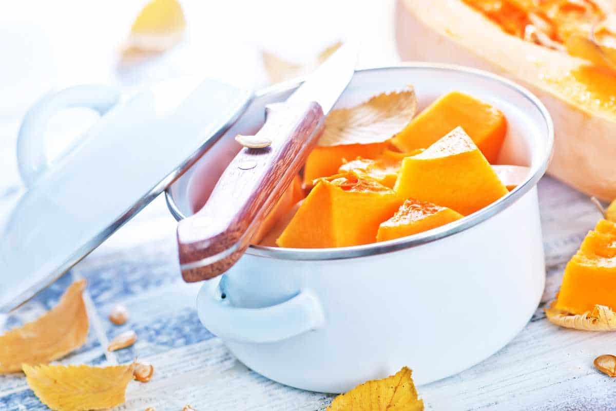 A white pot filled with cubed pumpkin pieces, topped with a wooden-handled knife, sits on a light surface surrounded by autumn leaves.