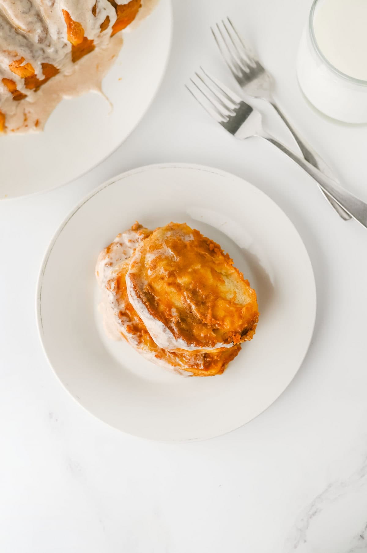 A pull apart pumpkin bread with icing on a white plate, next to two forks and a glass of milk on a marble surface.