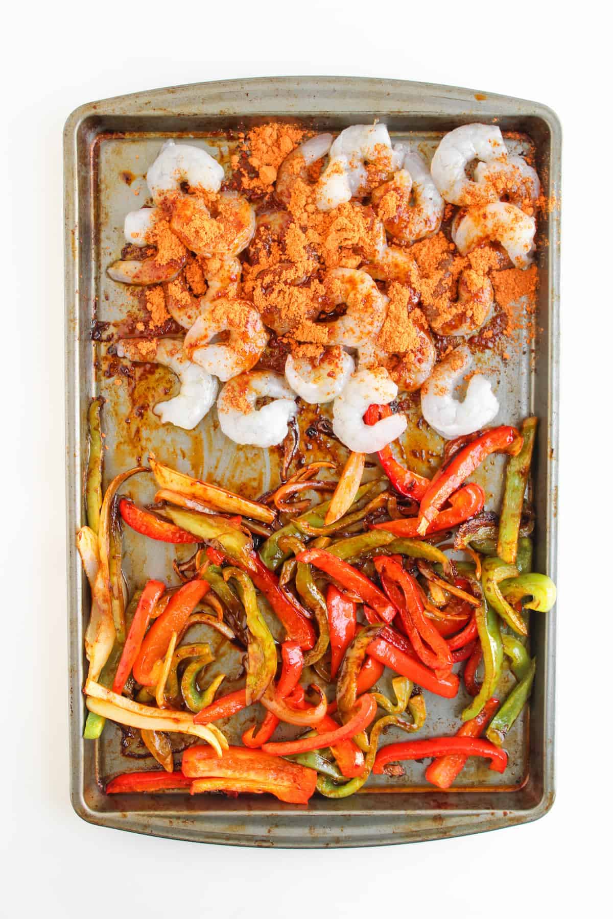 A baking sheet with seasoned raw shrimp on one side and cooked sliced red and green bell peppers on the other side.
