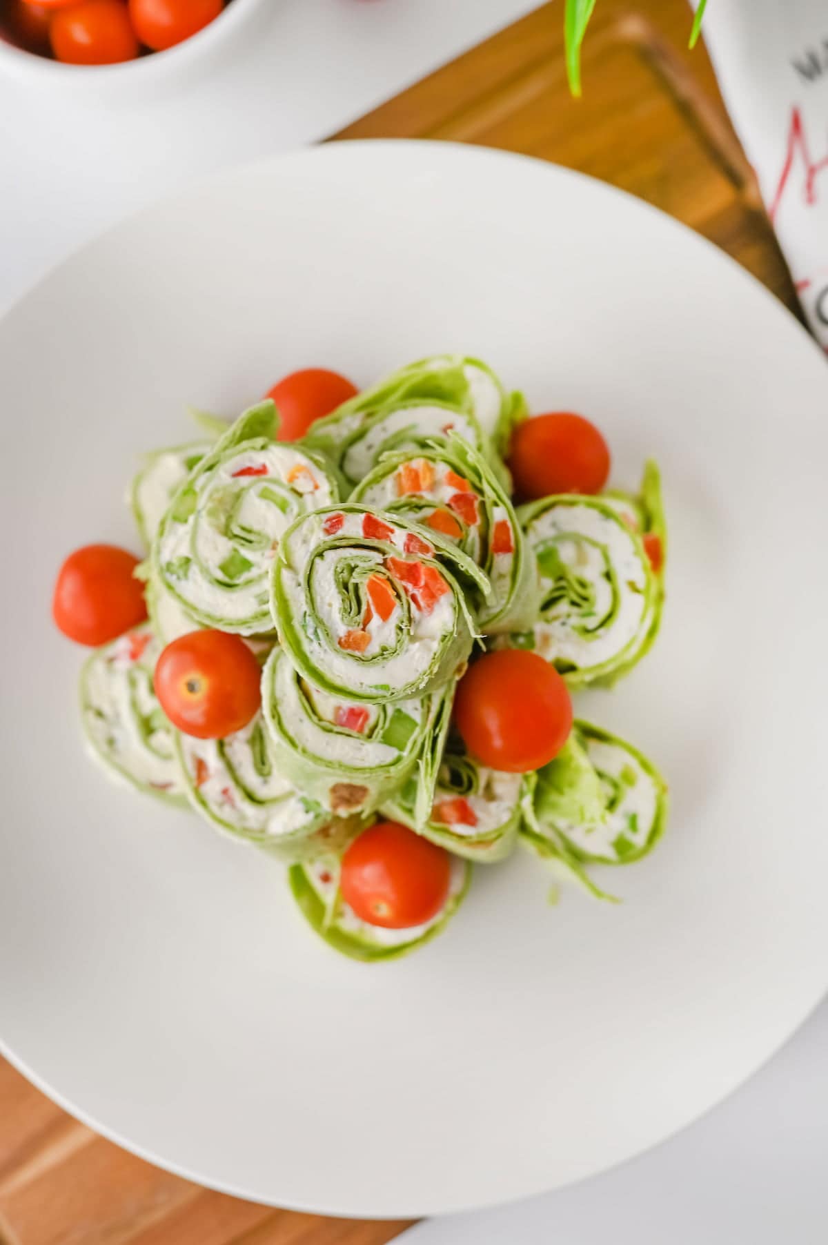 A white plate with spinach tortilla pinwheels filled with cream cheese and diced red peppers, garnished with whole cherry tomatoes.
