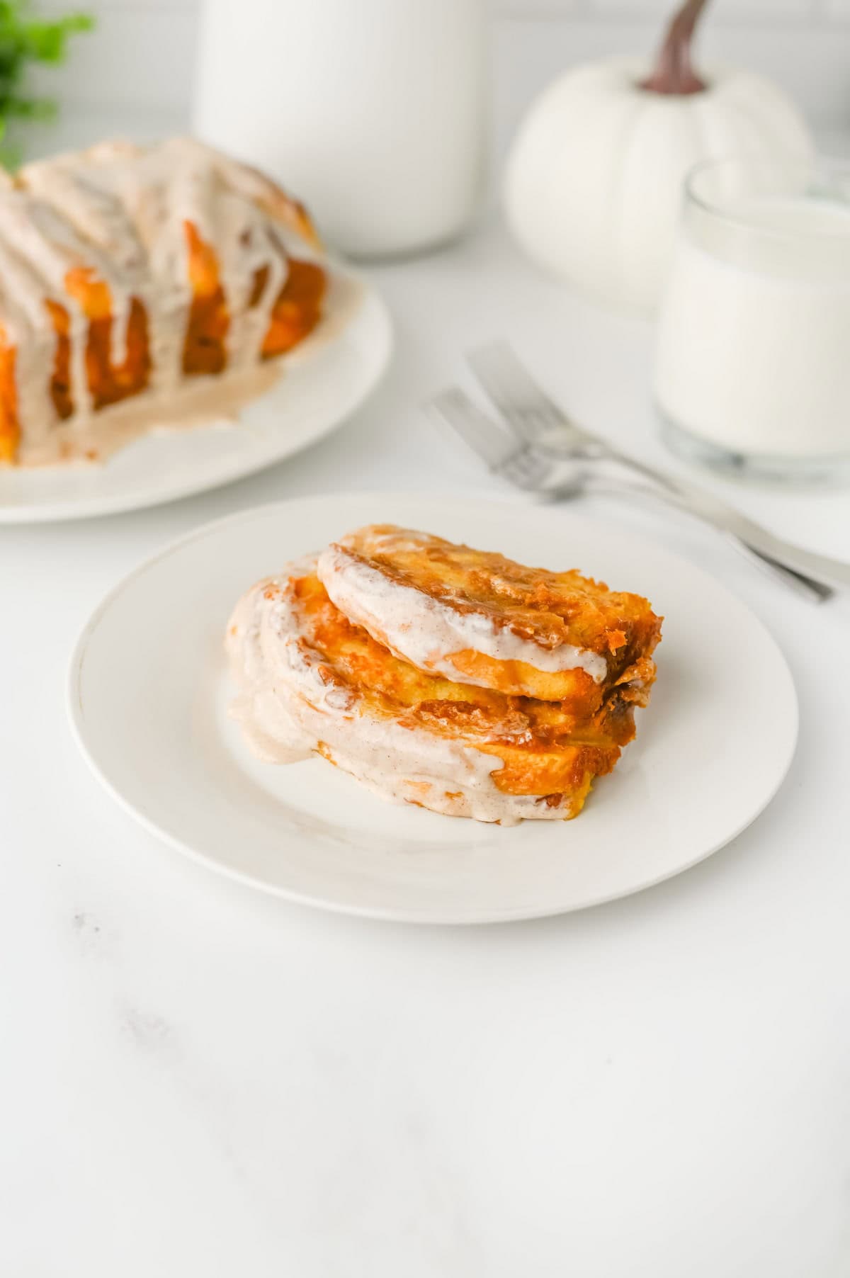 Two slices of pull-apart pumpkin bread with icing on a white plate, with a glass of milk and more bread in the background.