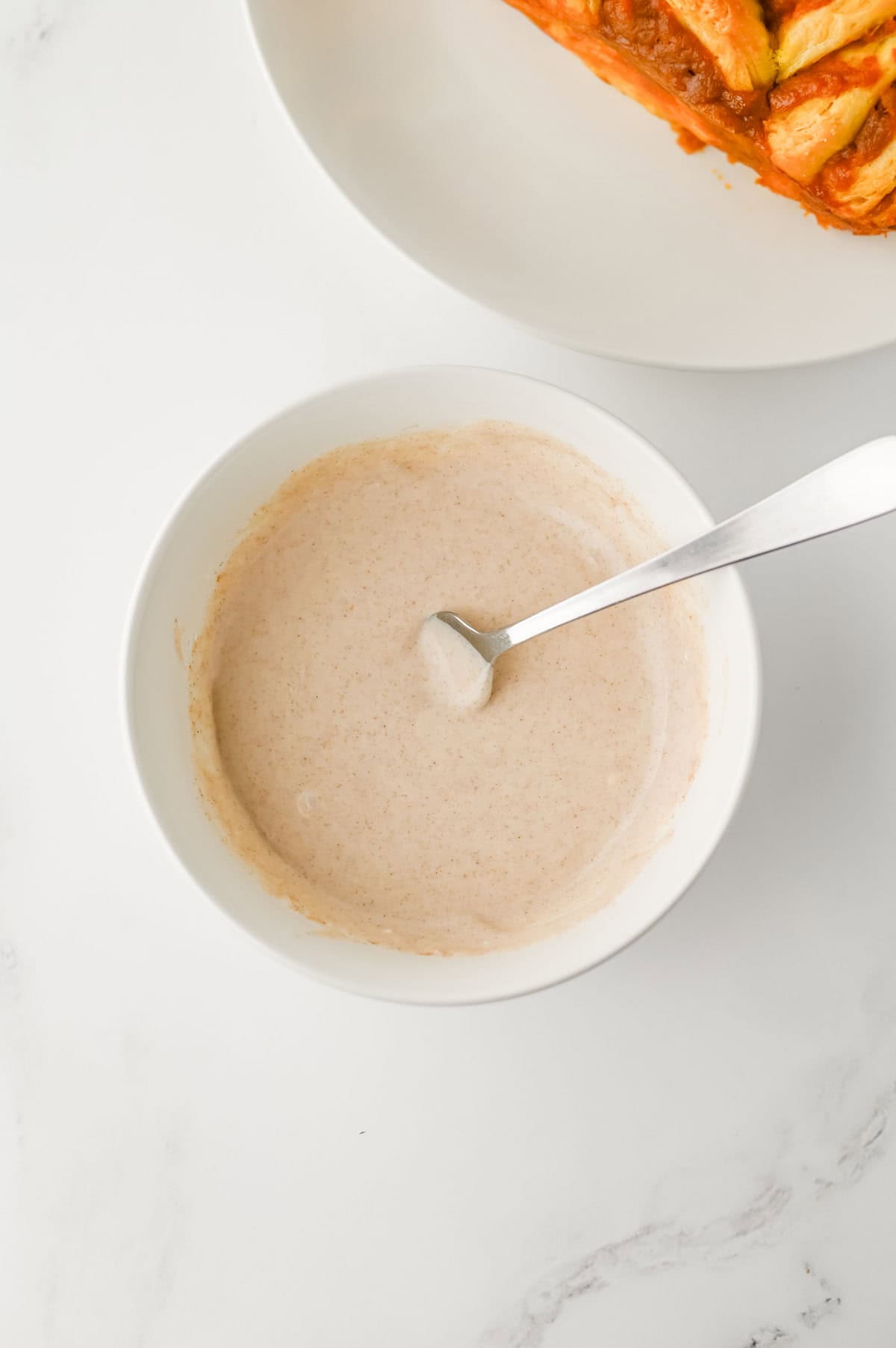 A white bowl containing a creamy, light brown mixture with a spoon inside, placed on a white surface next to a white plate with food partially visible.