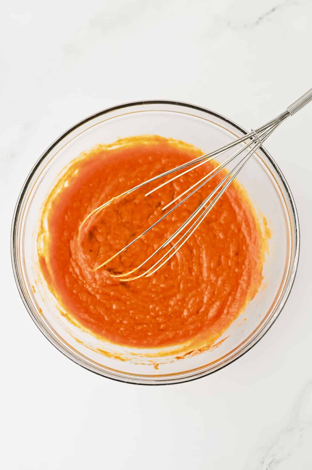 A glass bowl containing orange batter being mixed with a metal whisk on a white surface.