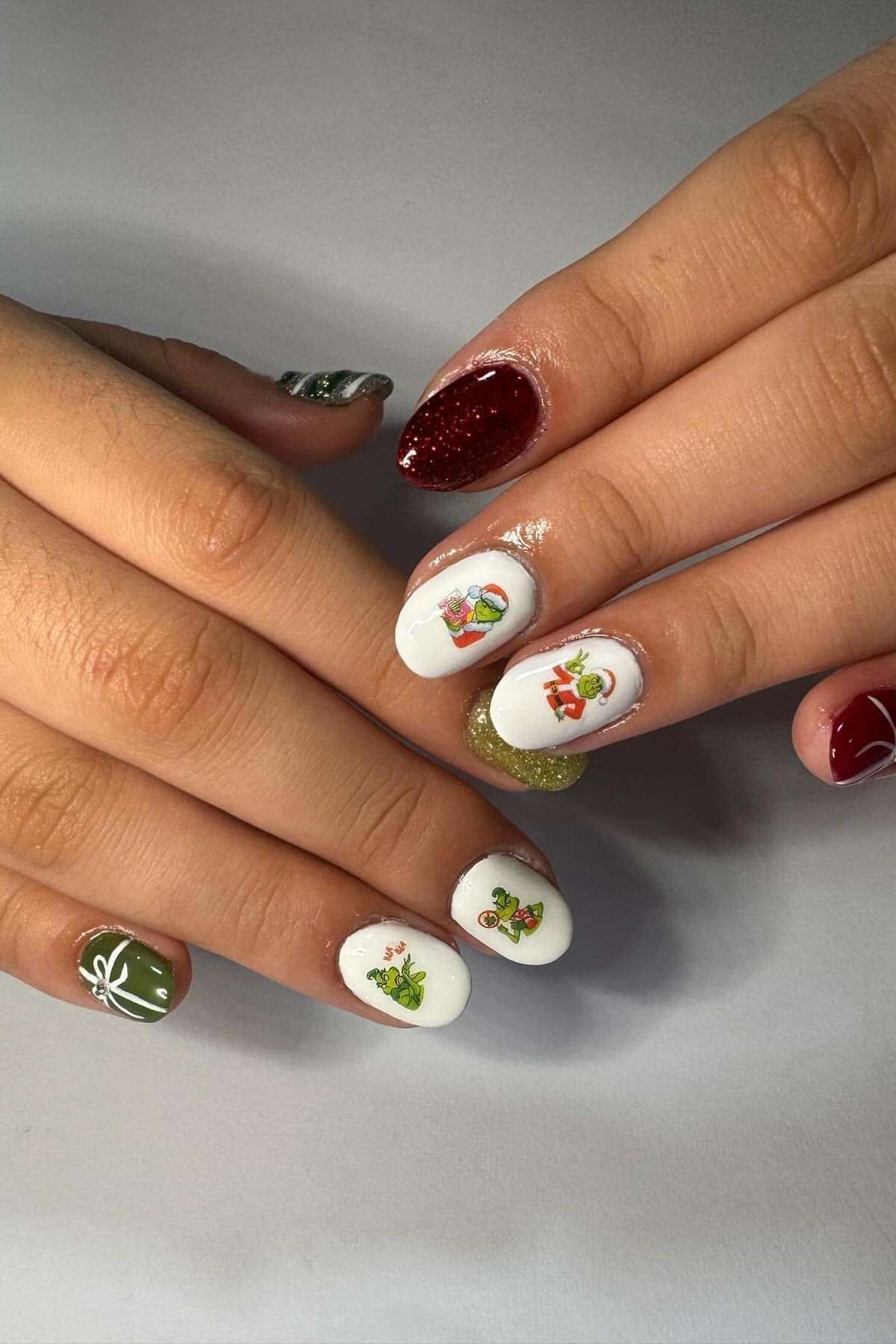 Hands with festive nail art featuring red, green, and white polish, glitter accents, and small Christmas-themed designs like presents and holly on a neutral background.