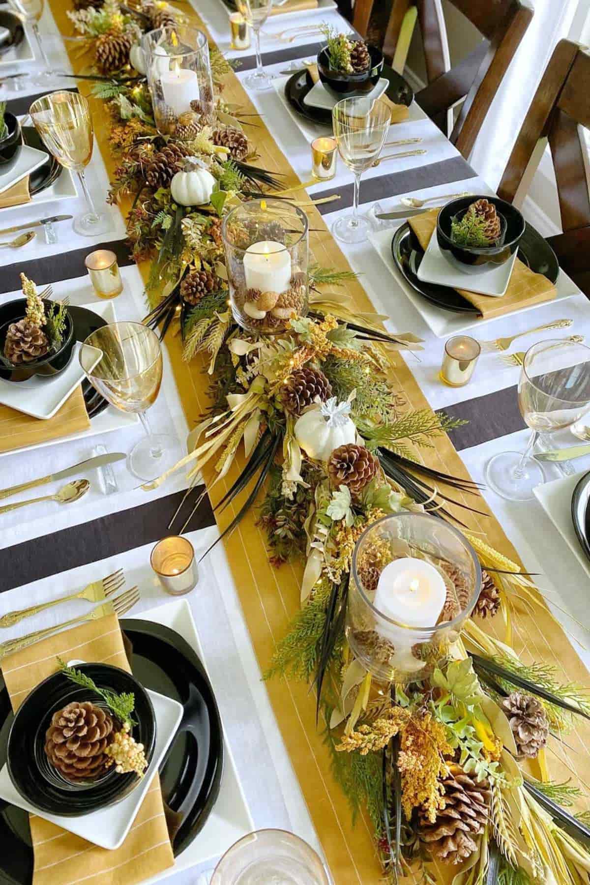 A long dining table set for a meal with black plates, gold napkins, wine glasses, pinecone decorations, and a centerpiece of candles, greenery, pinecones, and small white pumpkins.