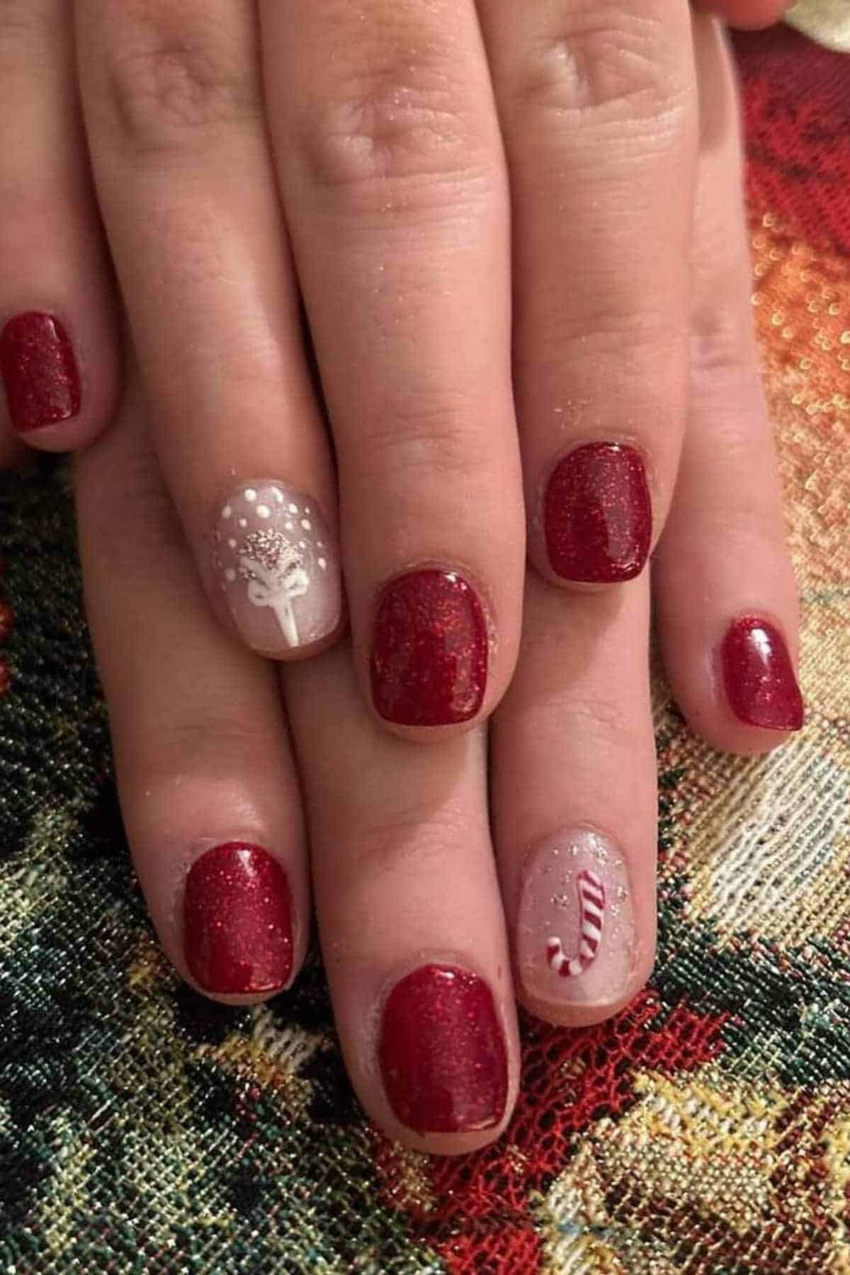 Close-up of hands with short, glittery red nails; ring fingers feature festive designs—one with a white holiday tree, the other with a red and white candy cane.