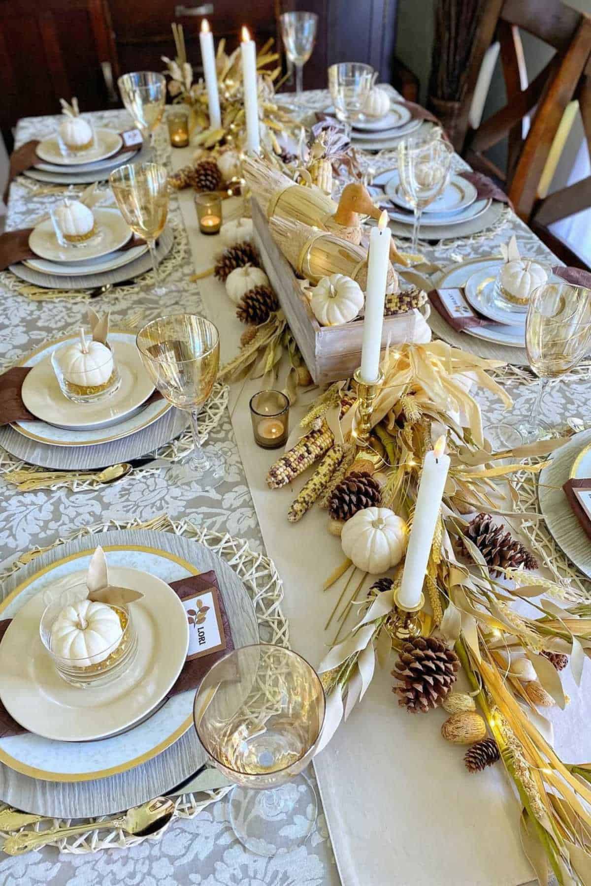 A fall-themed dining table set with plates, gold-rimmed glasses, candles, mini white pumpkins, pine cones, dried corn, and foliage as centerpiece decorations.