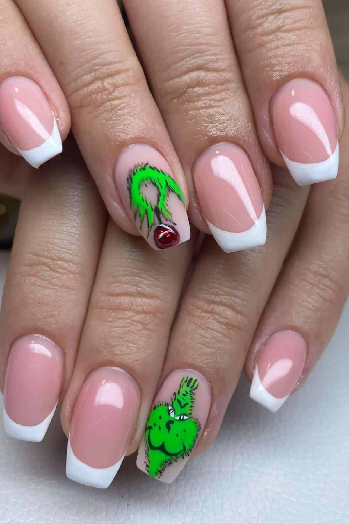 Hands with a French manicure; ring fingers feature green Grinch nail art with one holding a red ornament and the other showing the Grinch's face.