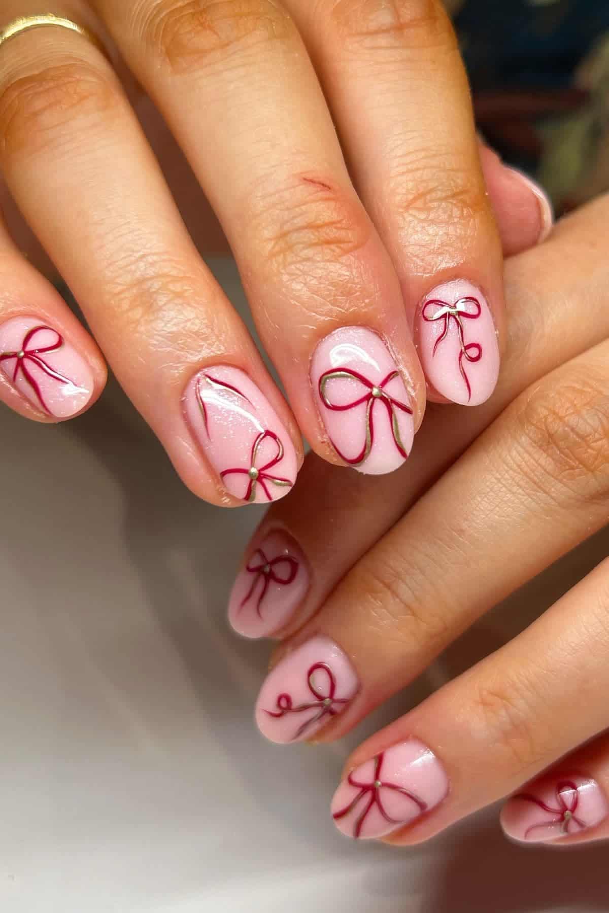 Close-up of hands with short, almond-shaped nails painted light pink and decorated with red bow designs on each nail.