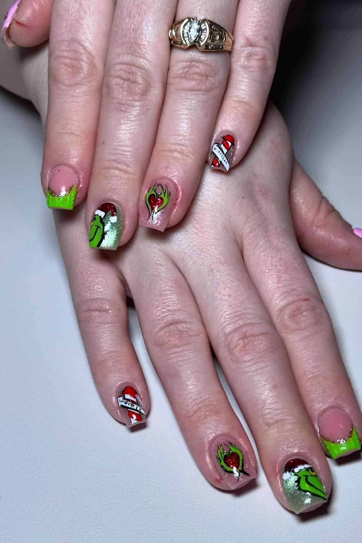 Hands with manicured nails featuring detailed nail art of The Grinch, green tips, hearts, and red and white accents. A gold ring is worn on one finger.