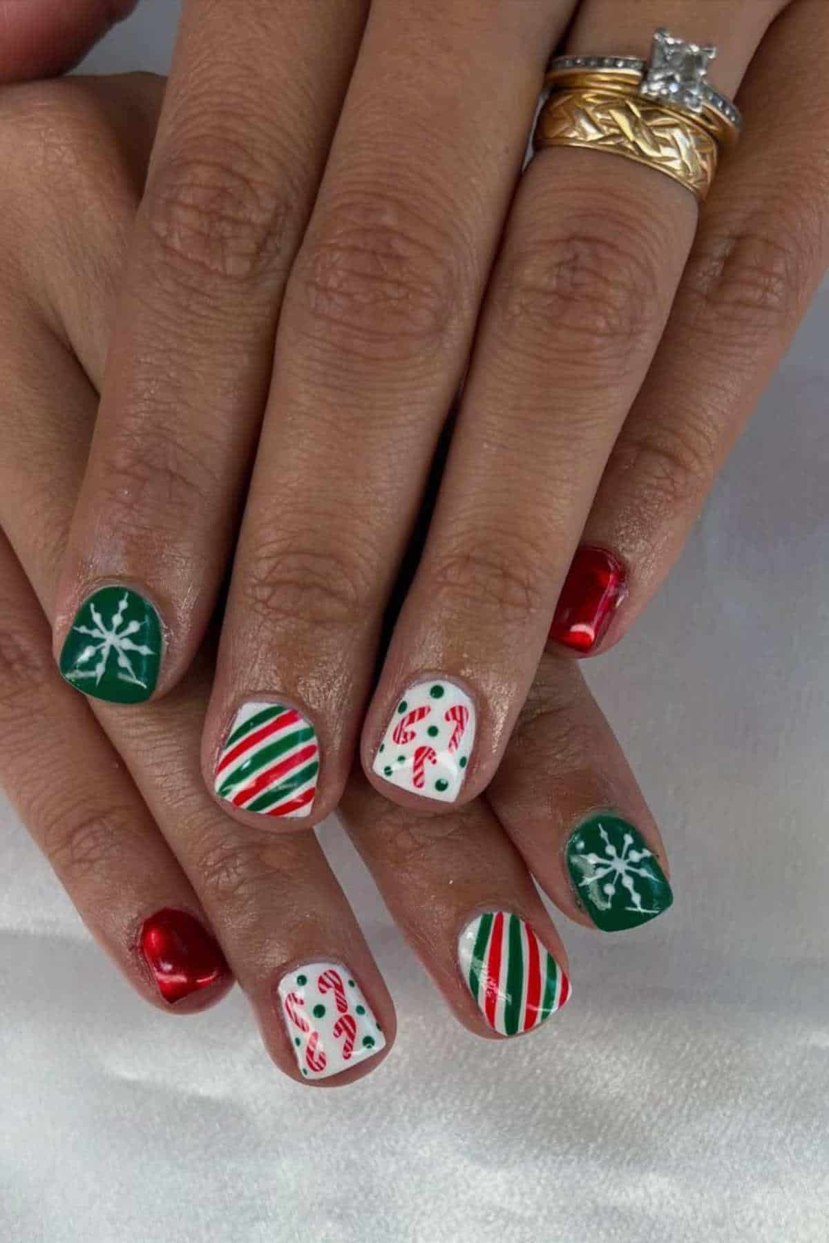 A hand with festive Christmas-themed nail art featuring candy canes, snowflakes, red and green stripes, and metallic red polish. The person is wearing gold and diamond rings.