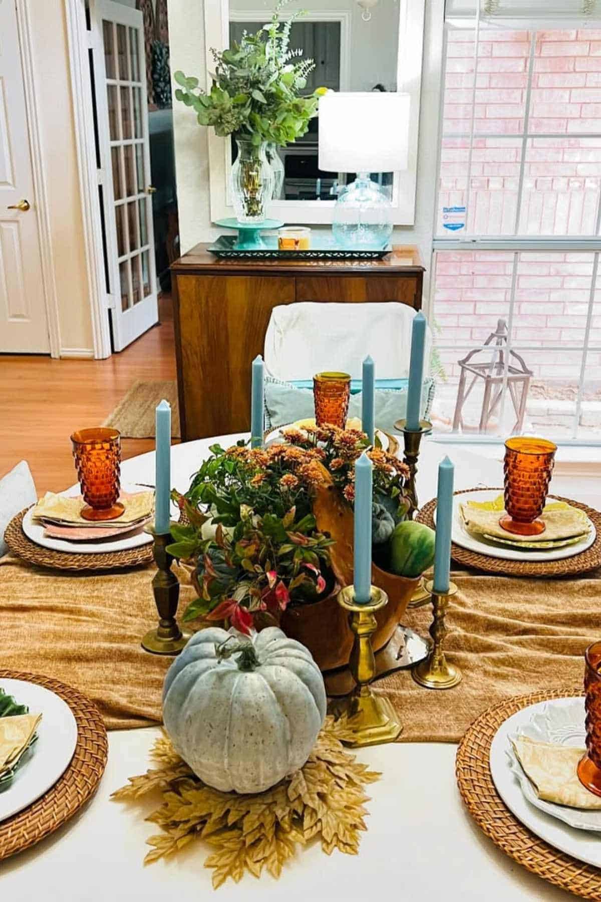 A dining table set with blue candles, amber glasses, a white pumpkin centerpiece, and a roasted turkey, with a lamp and vase on a sideboard in the background.