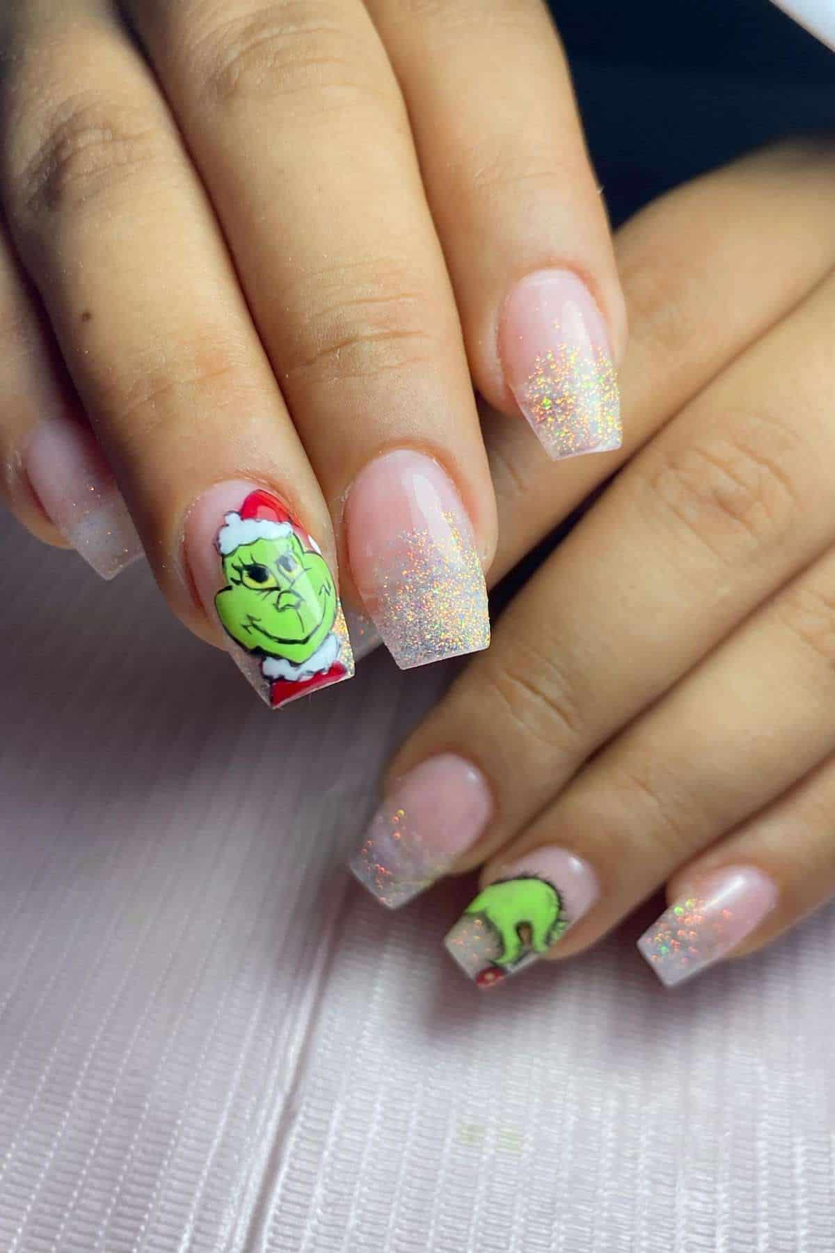 Hands with a manicure featuring glittery ombre nails and accent nails with hand-painted Grinch faces wearing Santa hats.